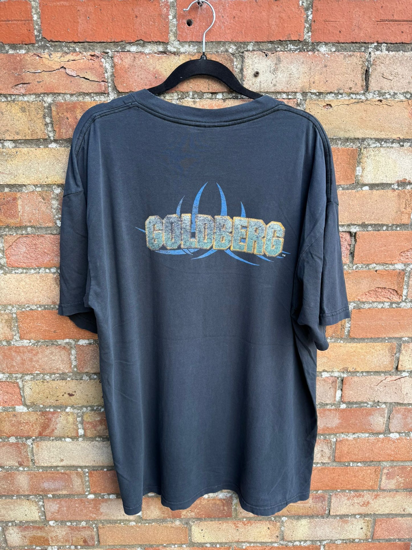 Back of a black t-shirt with Goldberg logo, hanging against a brick wall