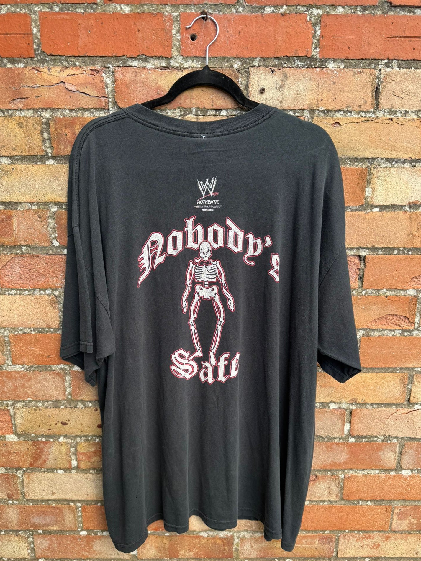 Back view of black t-shirt with skeleton white and red graphic design and Nobody's Safe text, on a hanger against a brick wall