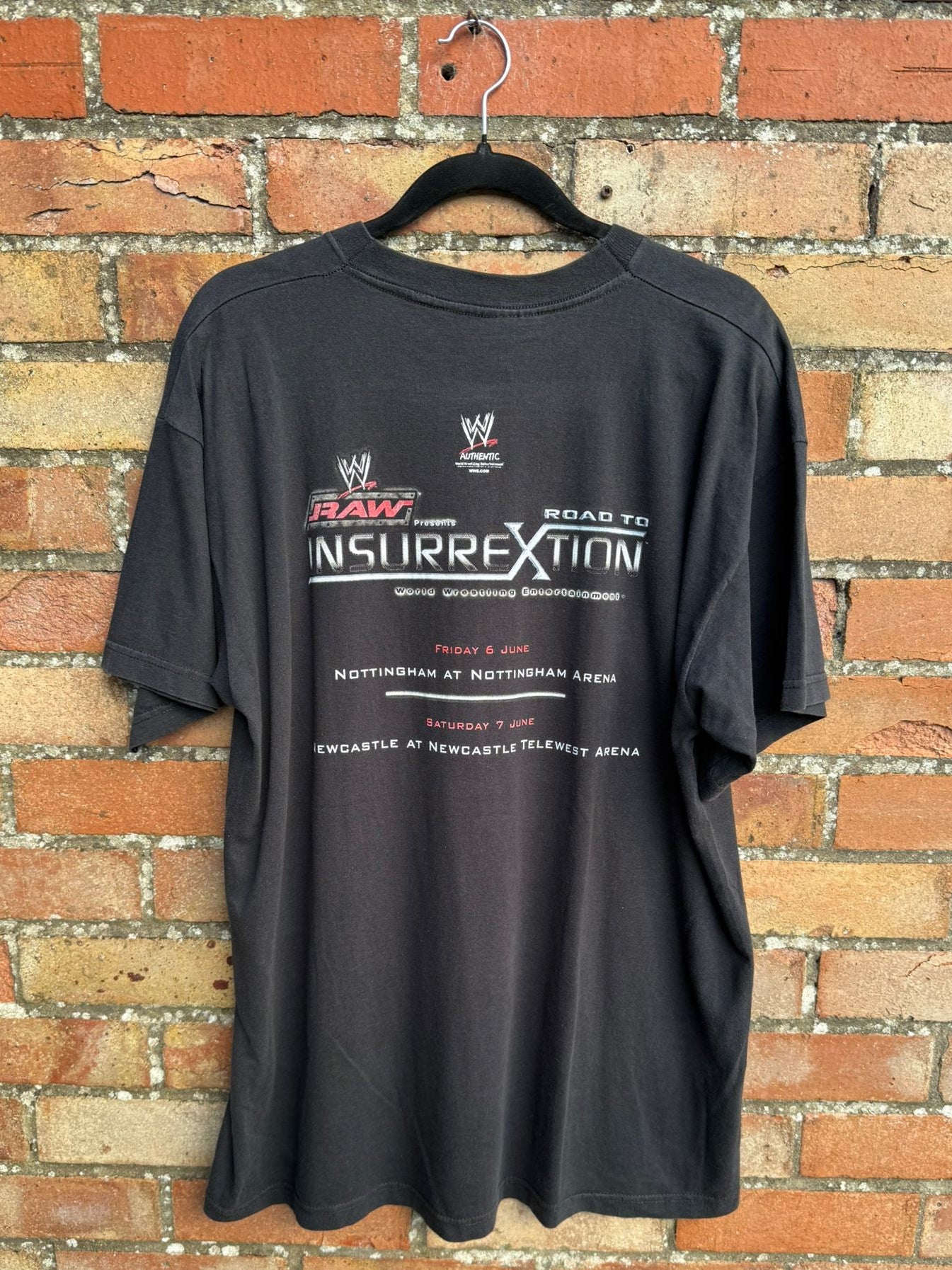 Back view of black t-shirt with wrestling event text on a hanger against a brick wall