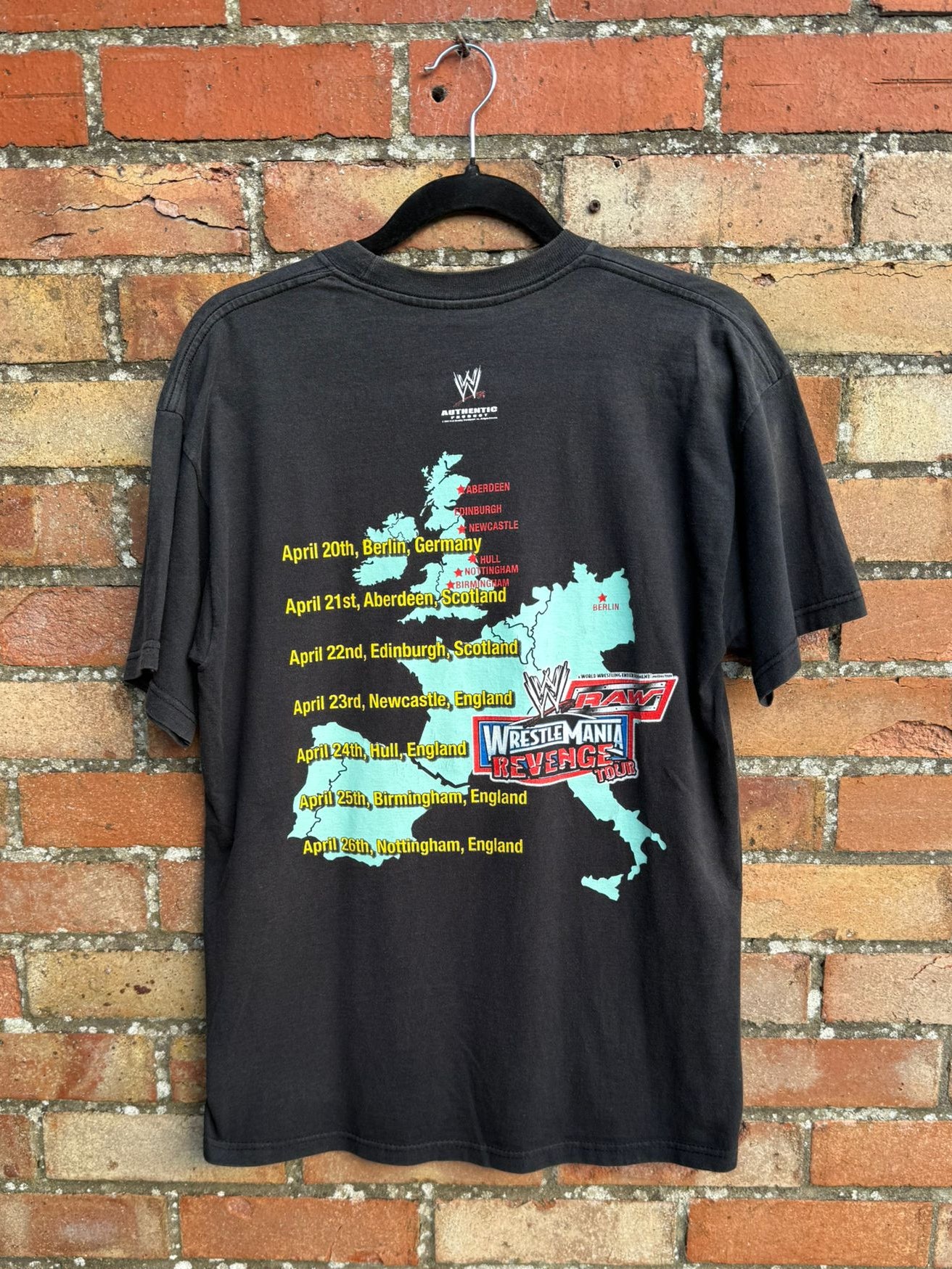 Back view of a black t-shirt with event map design on a hanger against a brick wall.