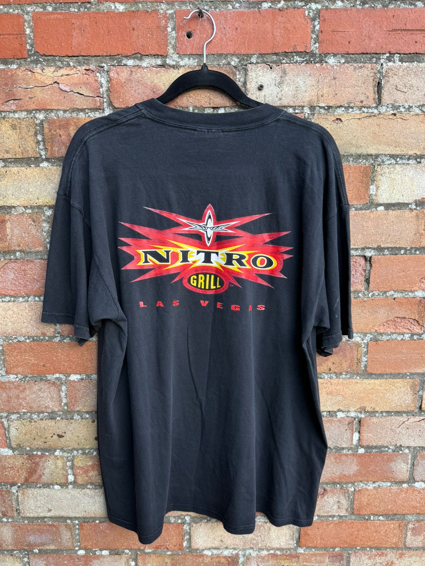 Black t-shirt with 'Nitro Grill Las Vegas' logo on a hanger against a brick wall.