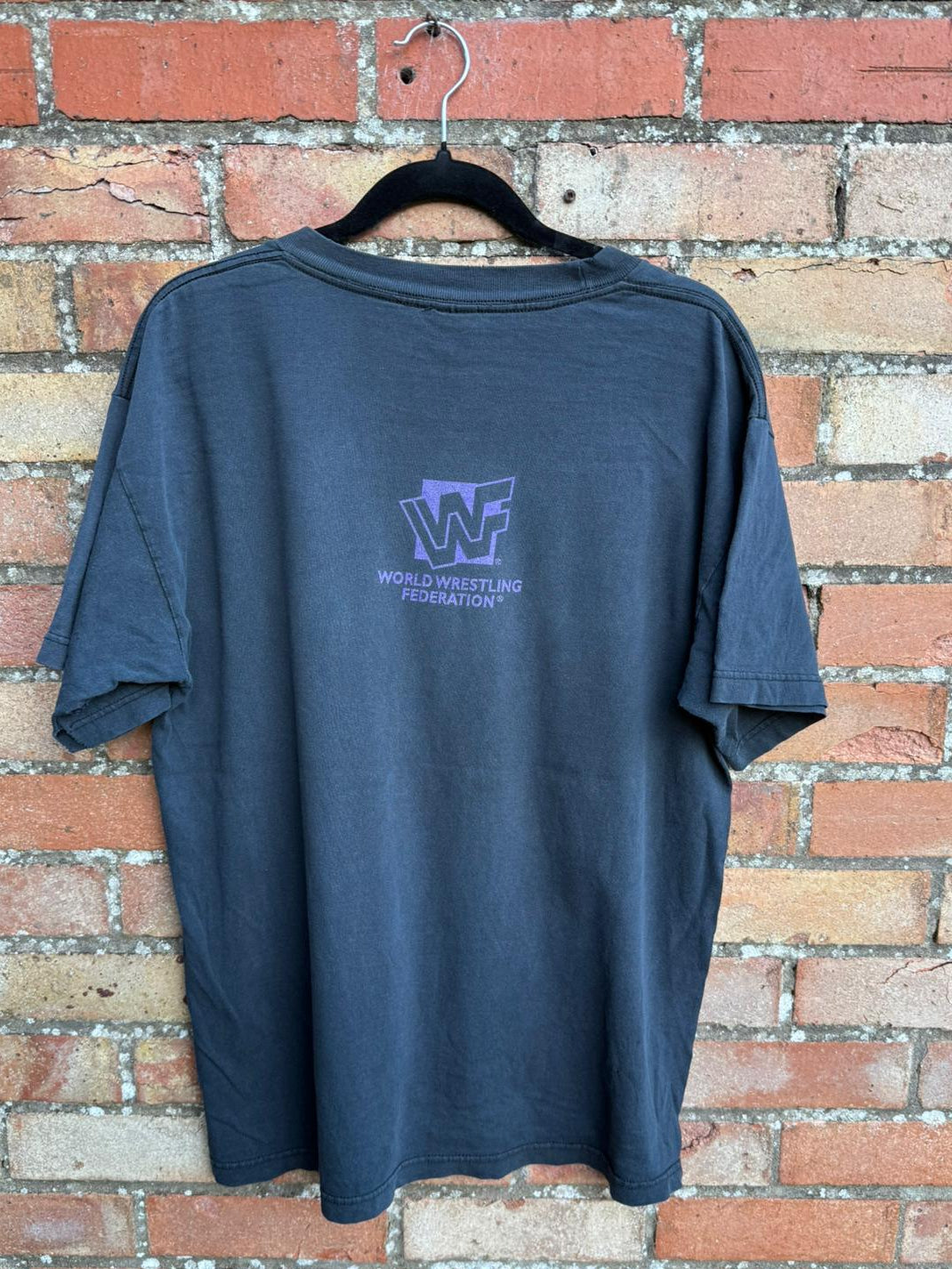 Black t-shirt with World Wrestling Federation logo on a hanger against a brick wall.