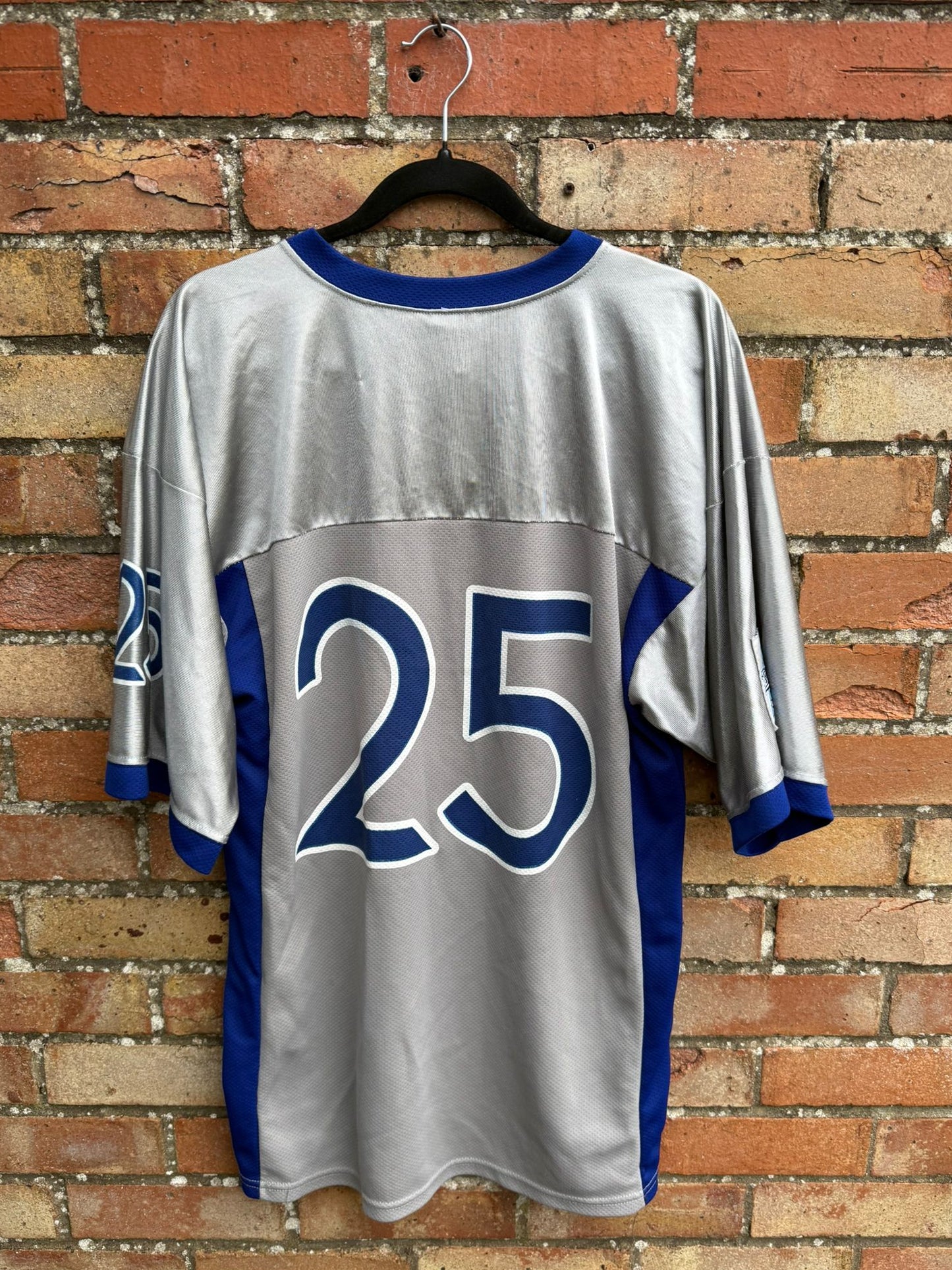 Back view of a silver sports jersey with blue number 25 on a hanger against a brick wall.