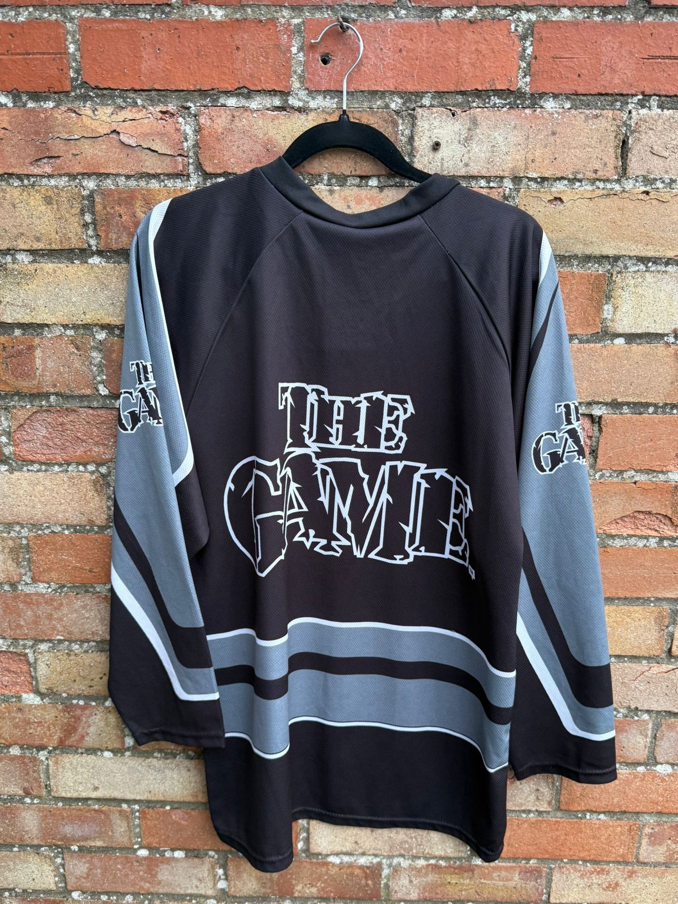 Bach view of a black and gray hockey-style jersey with 'The Game' text on a hanger against a brick wall.