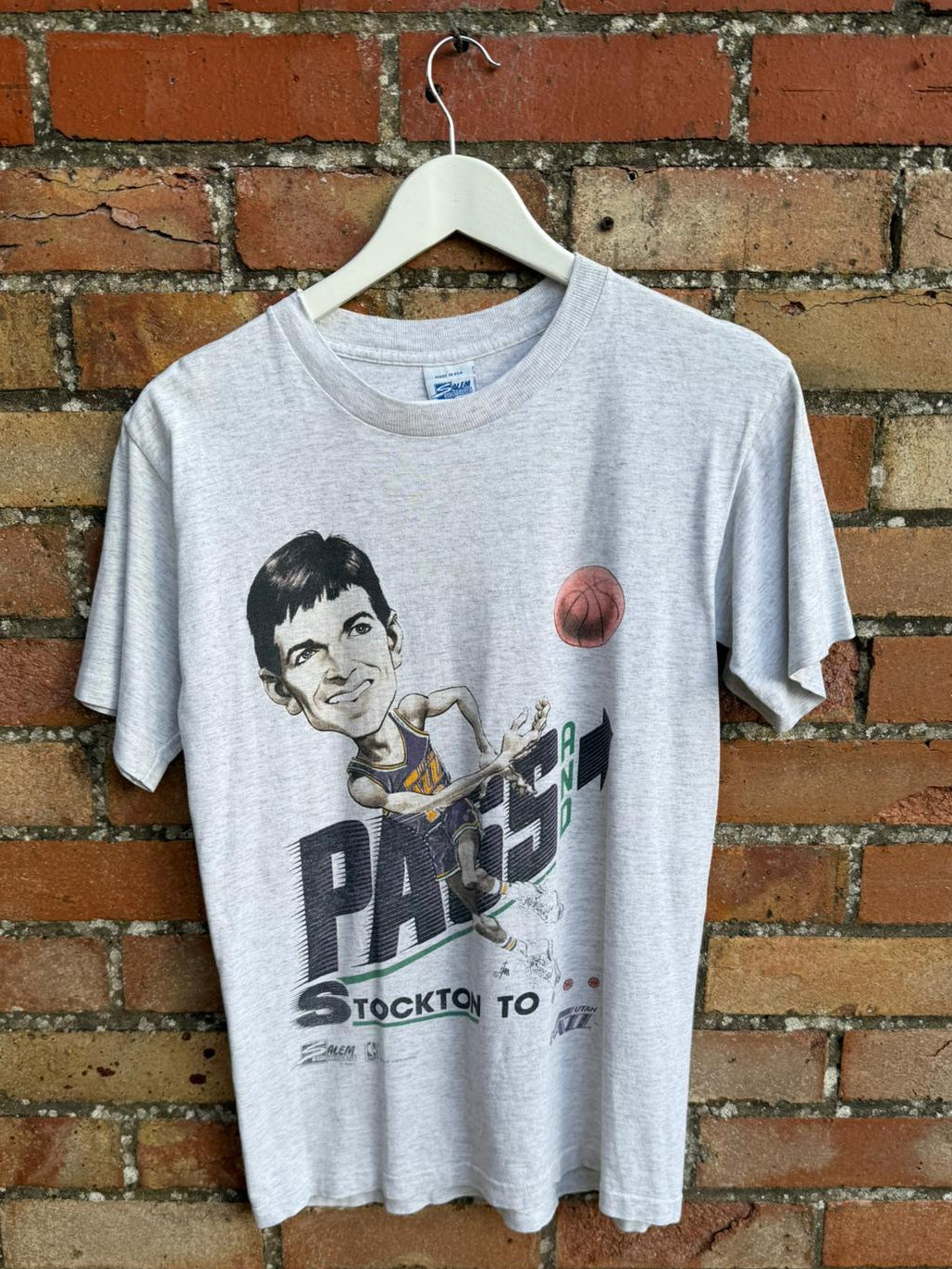 Gray t-shirt with Pass and Smash graphic design of a person playing basketball, hanging on a brick wall.