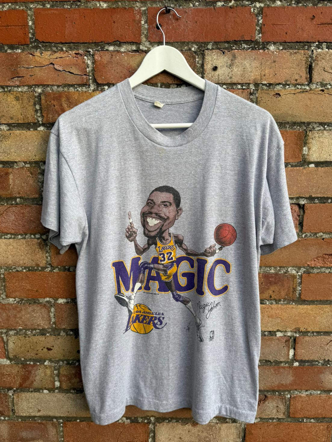 Gray t-shirt with a graphic of basketball Magic Johnson Los Angeles Lakers player, hanging on a brick wall.