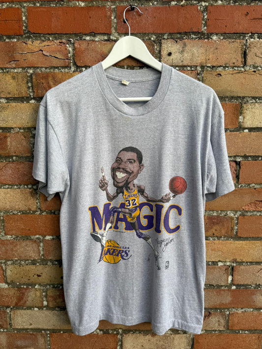 Gray t-shirt with a graphic of basketball Magic Johnson Los Angeles Lakers player, hanging on a brick wall.