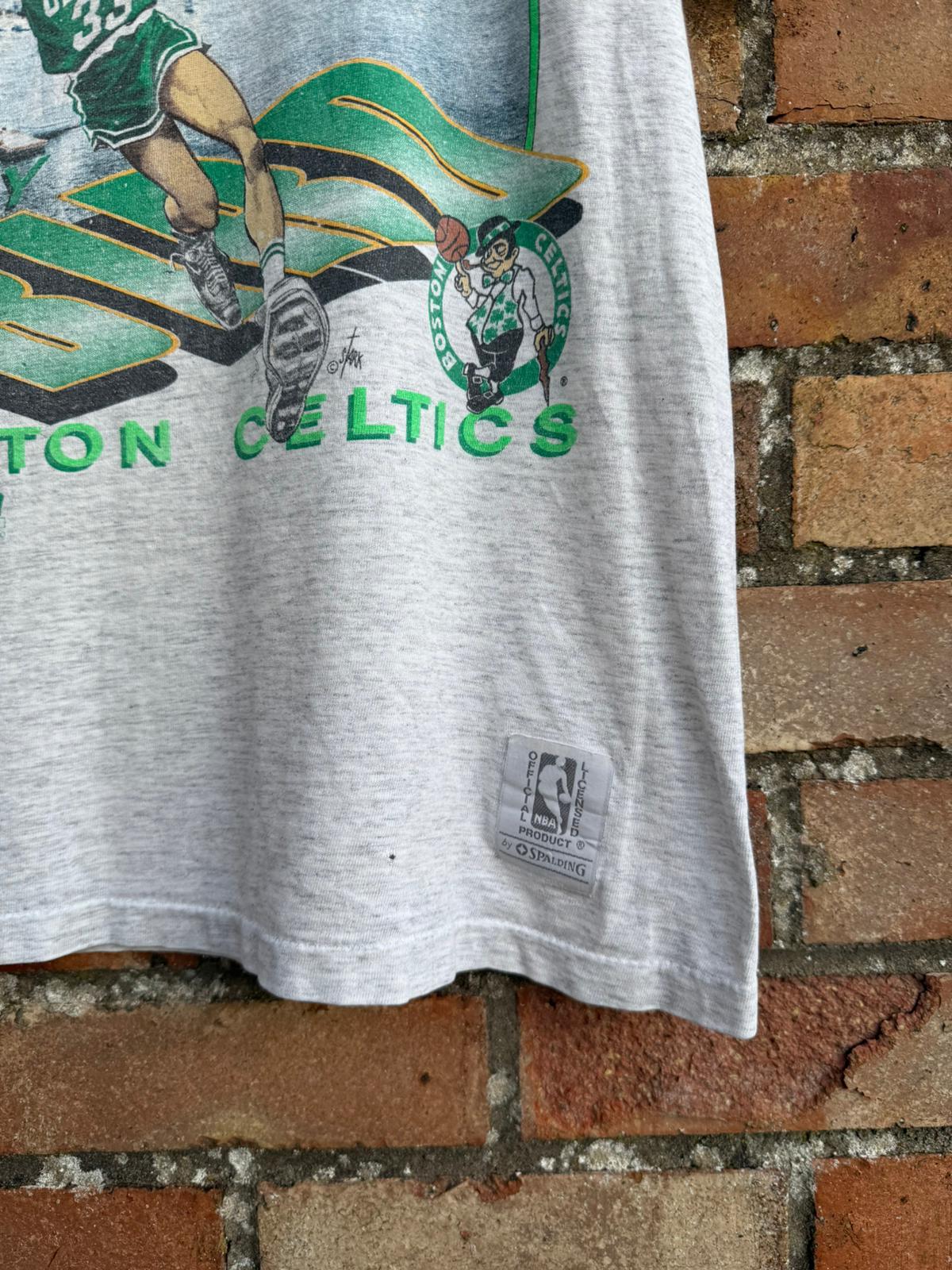 Gray t-shirt with Boston Celtics logo and Official NBA branding label, with a brick wall background