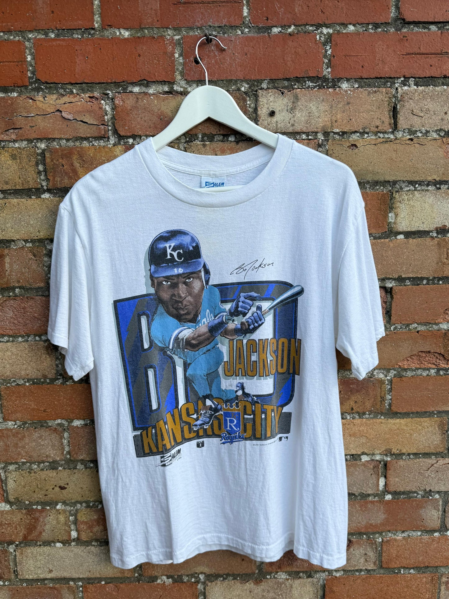 White t-shirt with a graphic of Kansas City baseball player Bo Jackson and text, hanging against a brick wall.