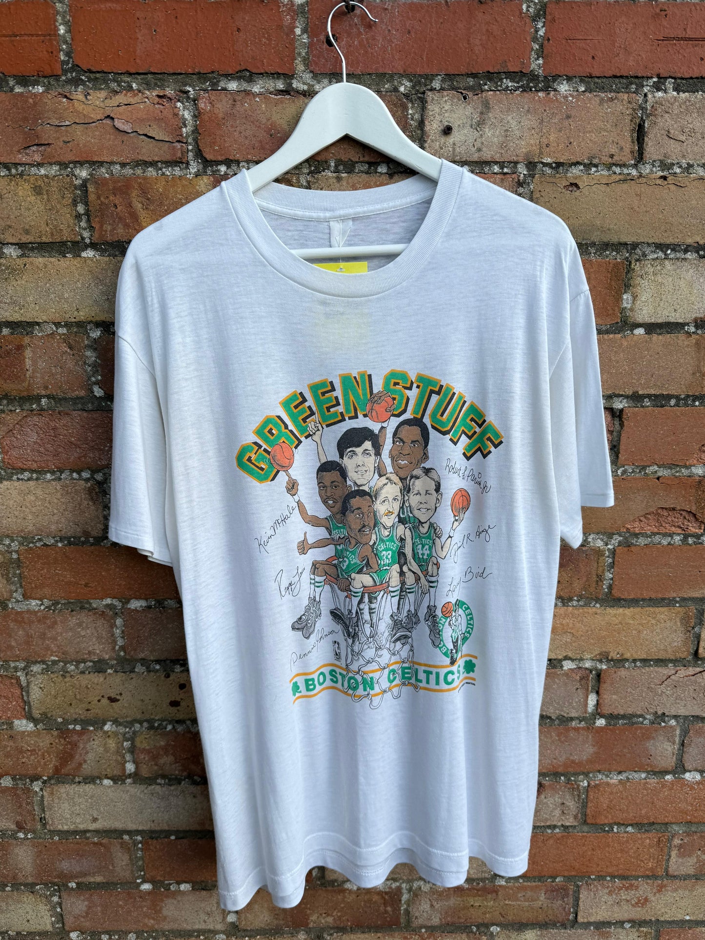 White t-shirt with text 'Green Stuff' and caricature of the Boston Celtics team including Larry Bird, hanging on a brick wall background