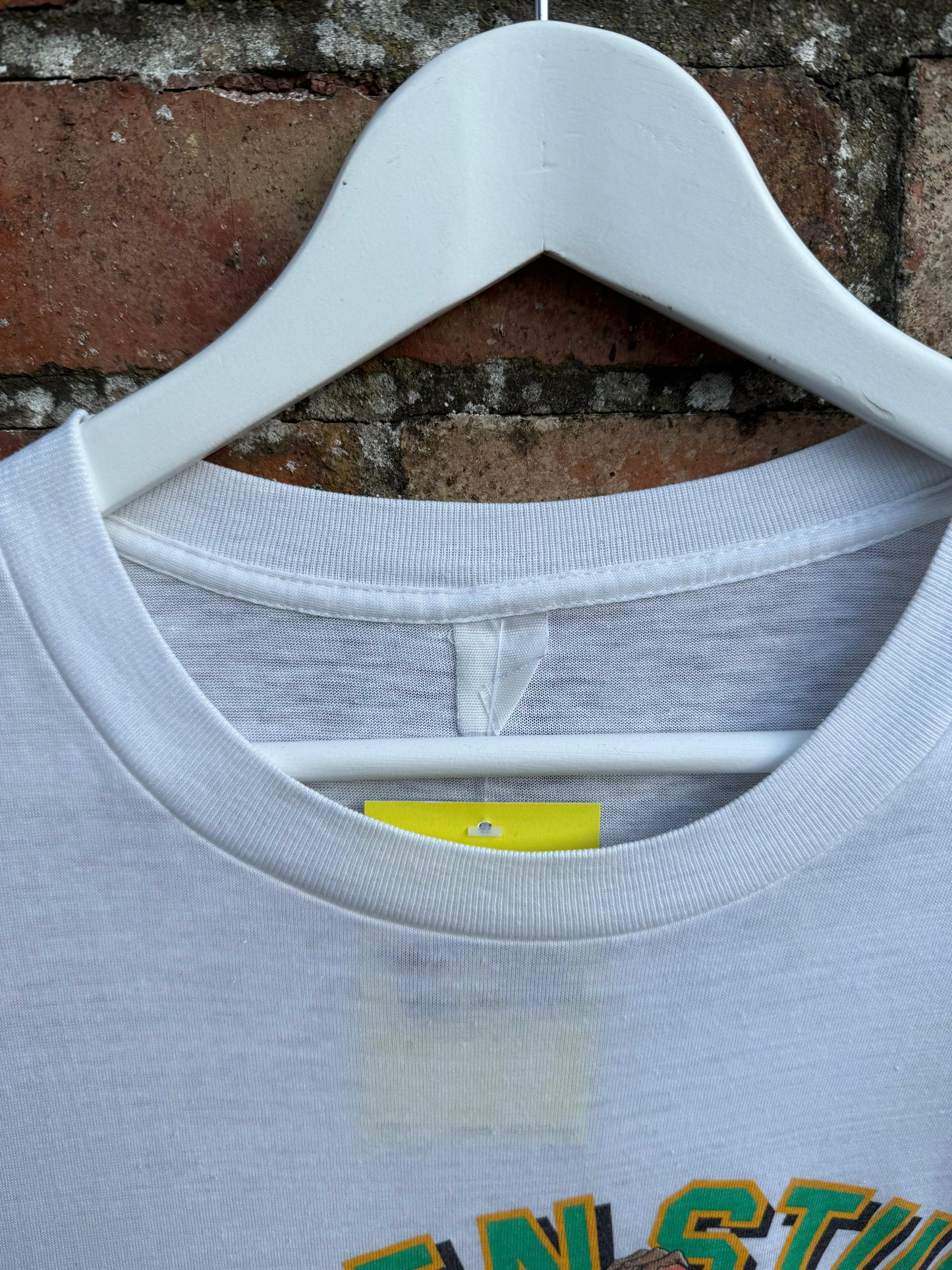 White t-shirt with a brand label on a hanger against a brick wall.