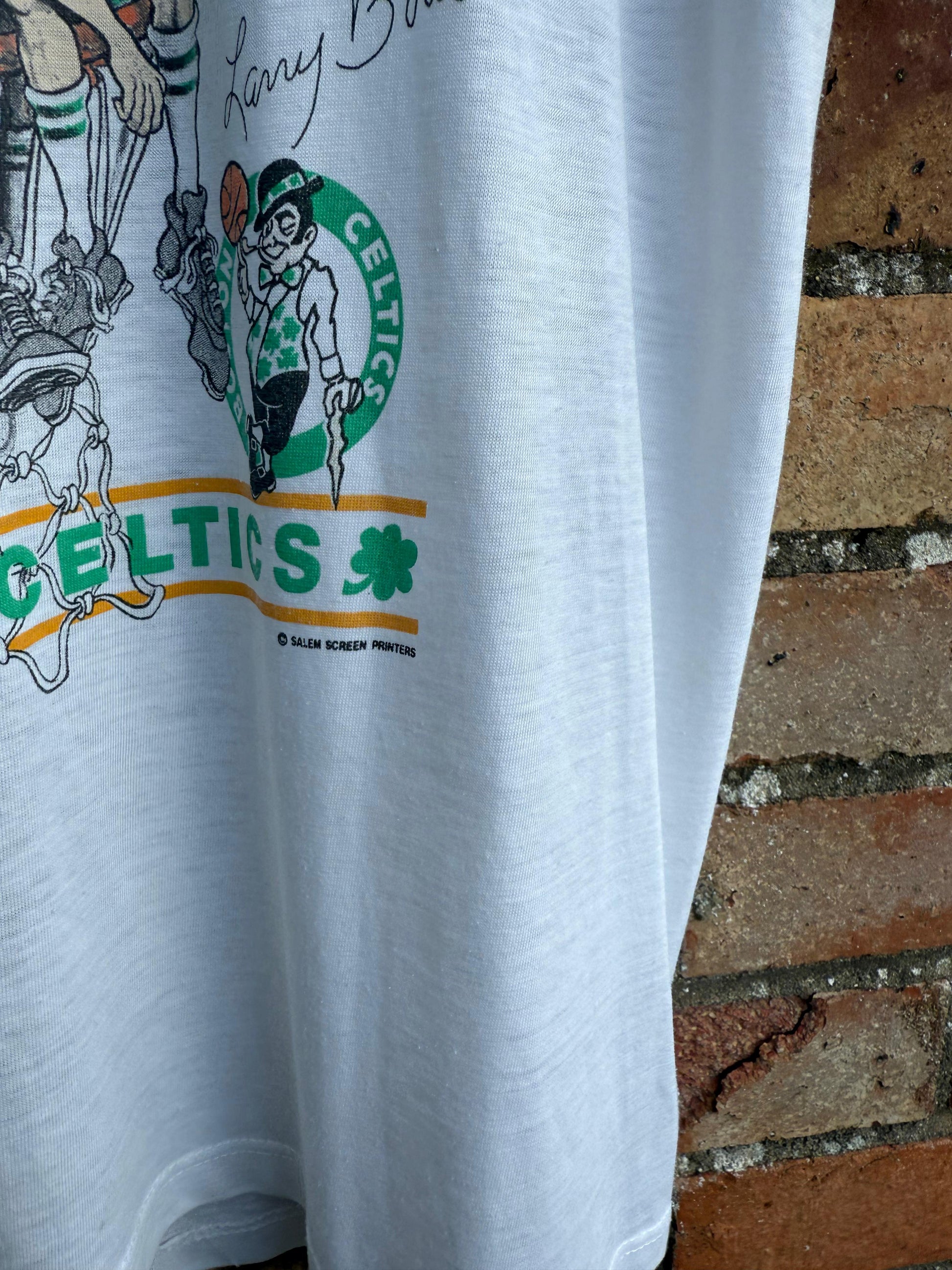 White t-shirt with Celtics logo and graphics, and Salem copyright, hanging on a brick wall background