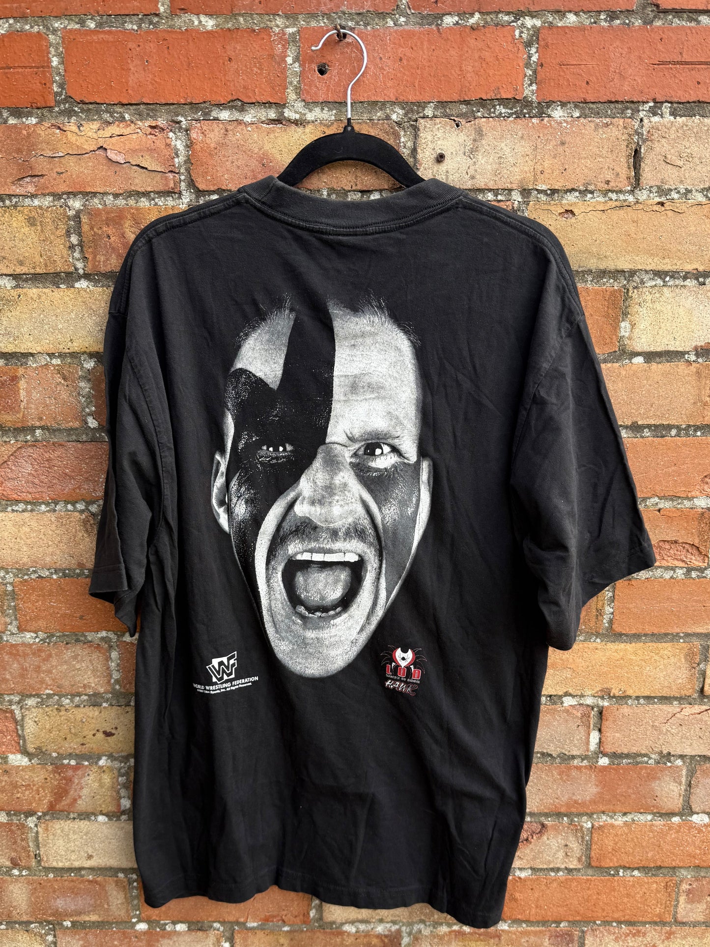 Black t-shirt with a face graphic design and LOD and WWF branding, hanging against a brick wall