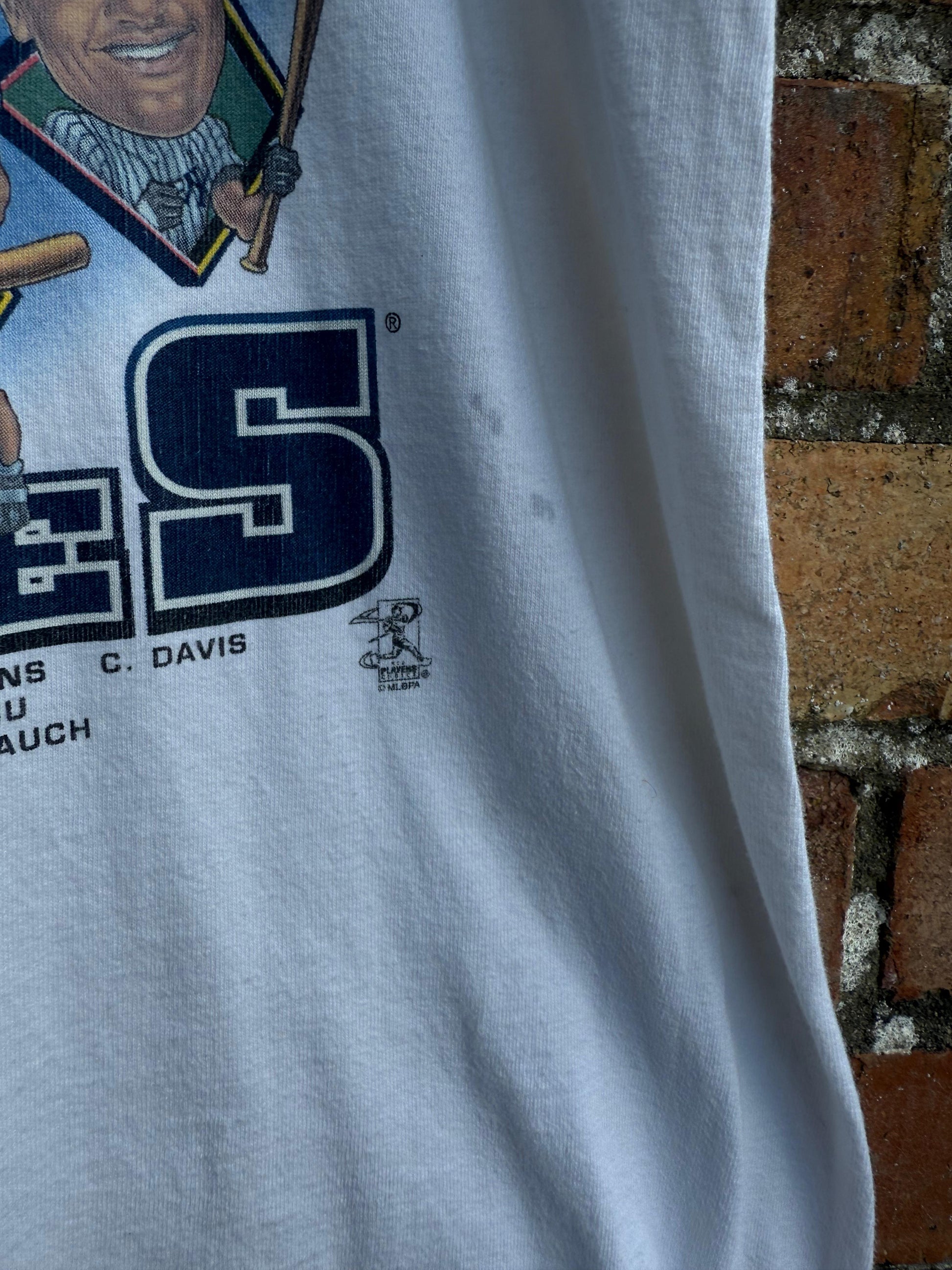 Close-up of a white t-shirt with a graphic design and text, showing a small area of staining, against a brick wall background.