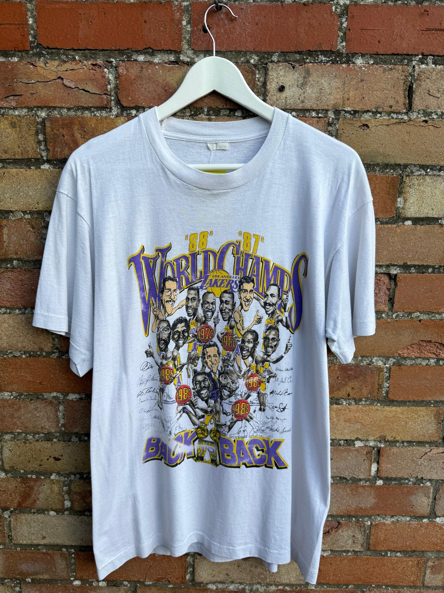White  Back to Back World Champs Lakers t-shirt with graphic design of basketball players and text, hanging against a brick wall.