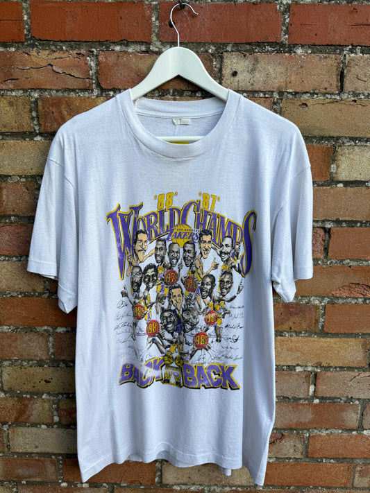 White  Back to Back World Champs Lakers t-shirt with graphic design of basketball players and text, hanging against a brick wall.