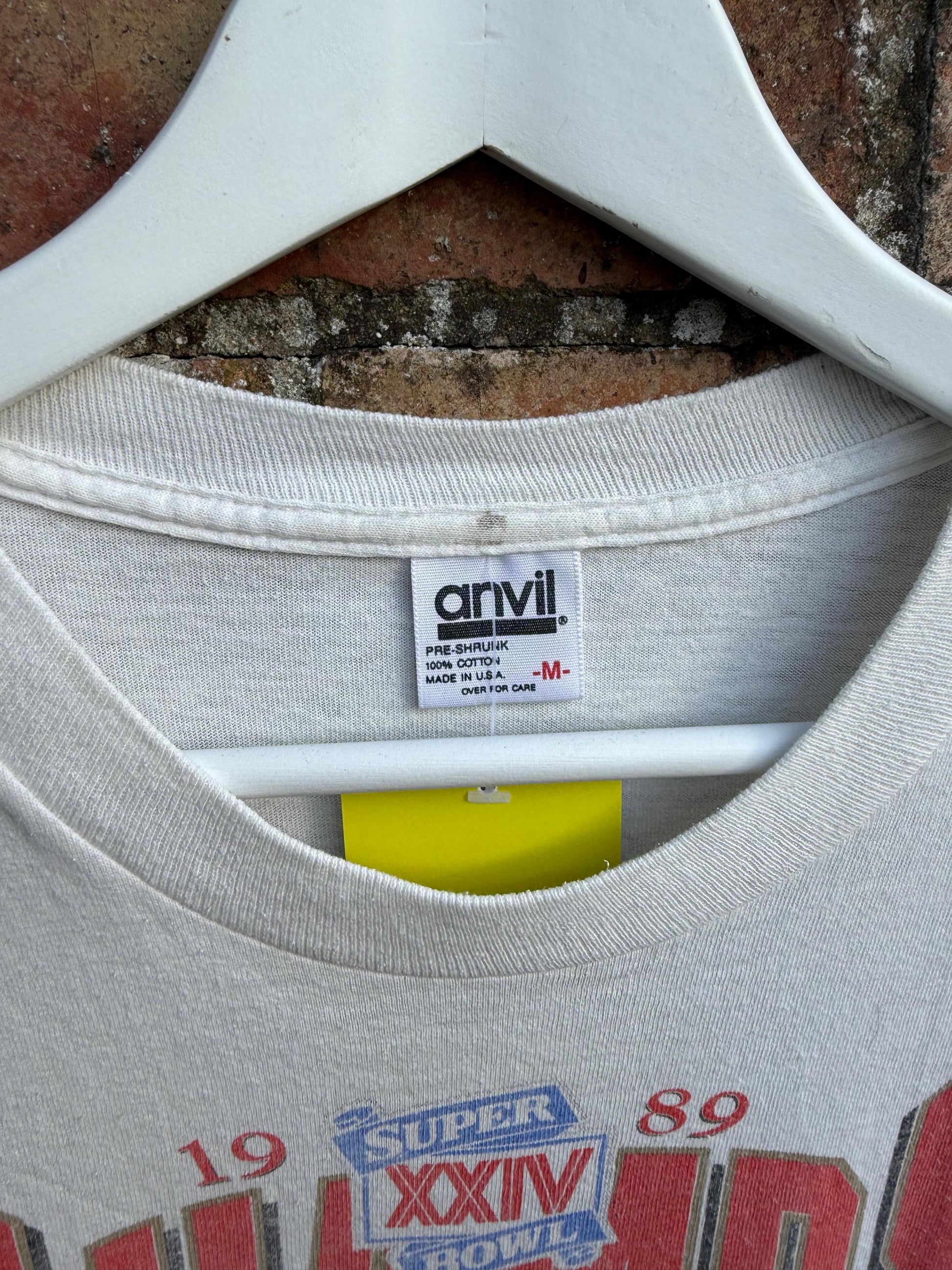 White t-shirt with 'Anvil' size M label and 1989 Super Bowl text on a hanger against a brick wall.