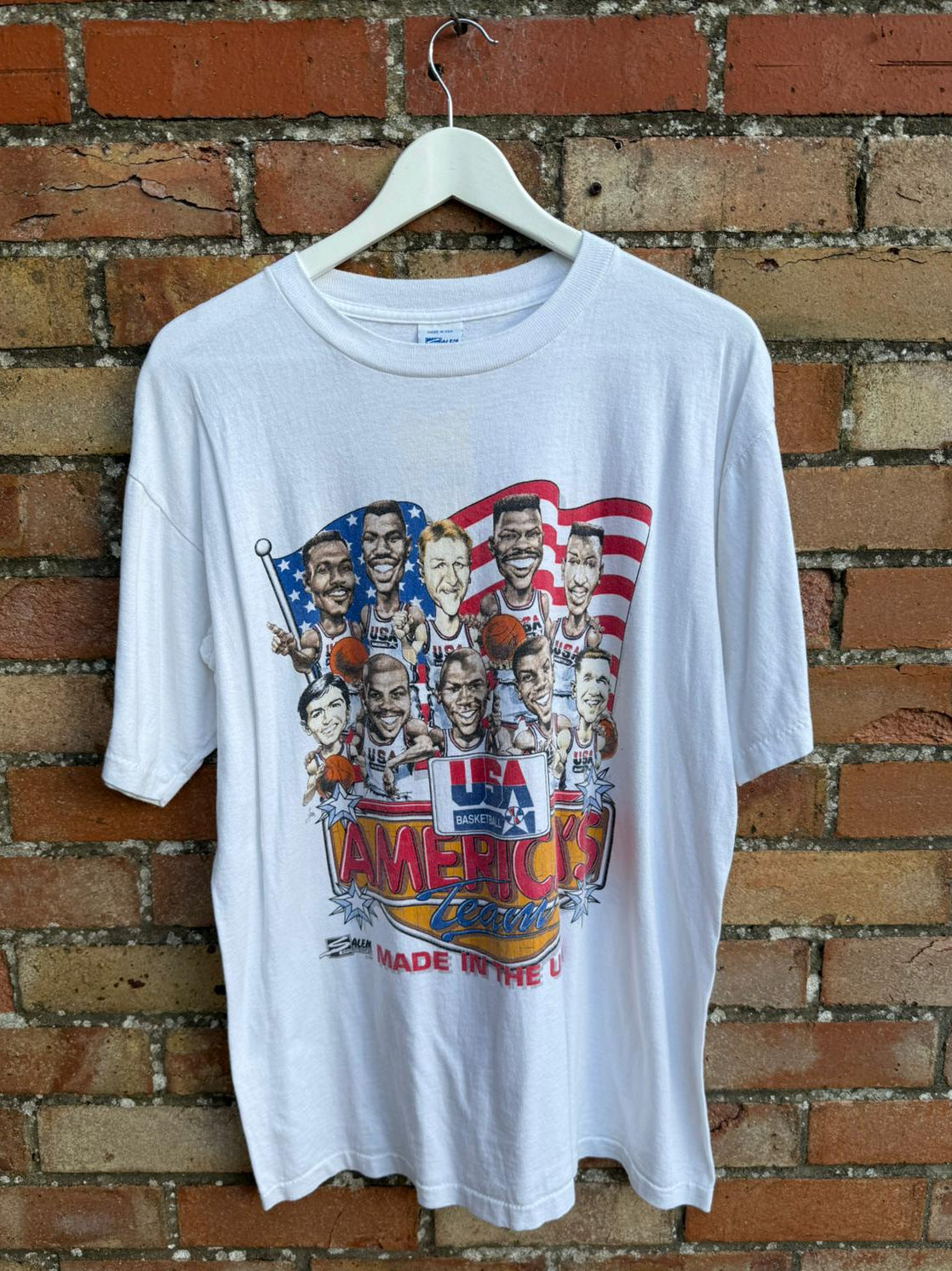 White t-shirt Made in USA America's Dream Team text with colorful graphic design on a hanger against a brick wall.