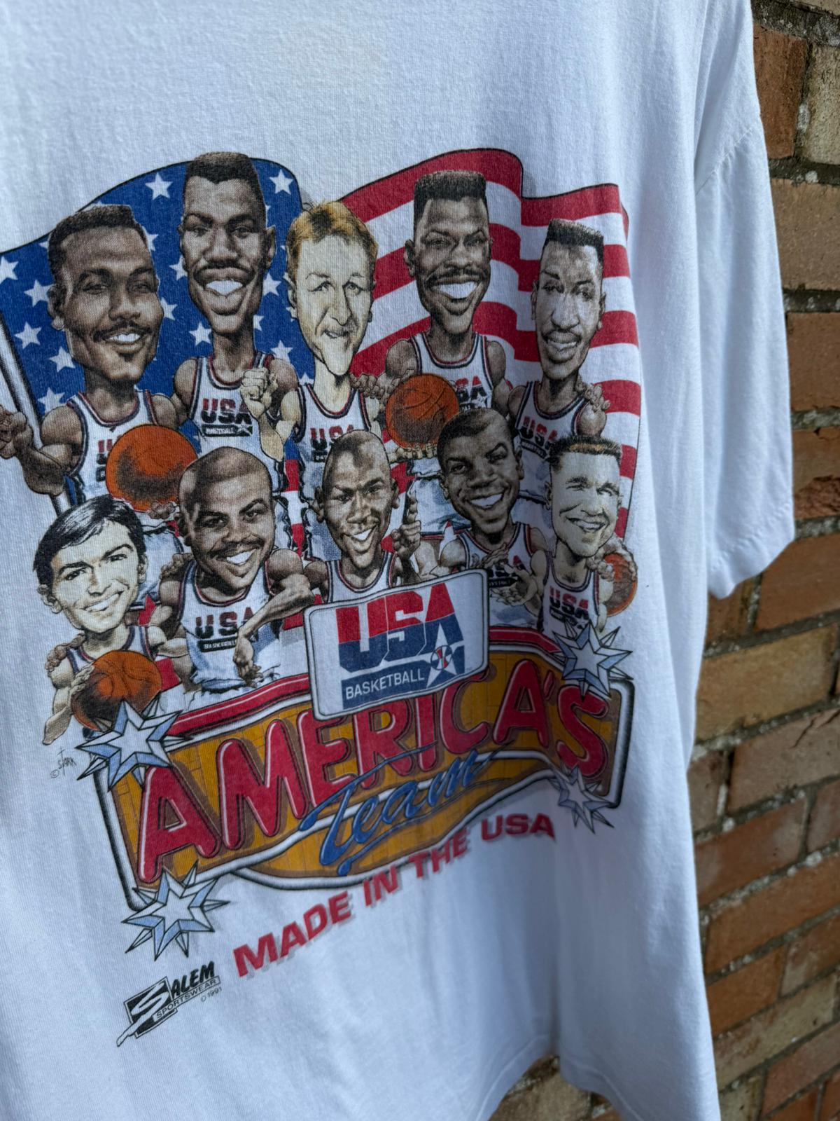 White t-shirt with a colorful graphic of basketball players and 'USA Basketball' text, hanging against a brick wall.
