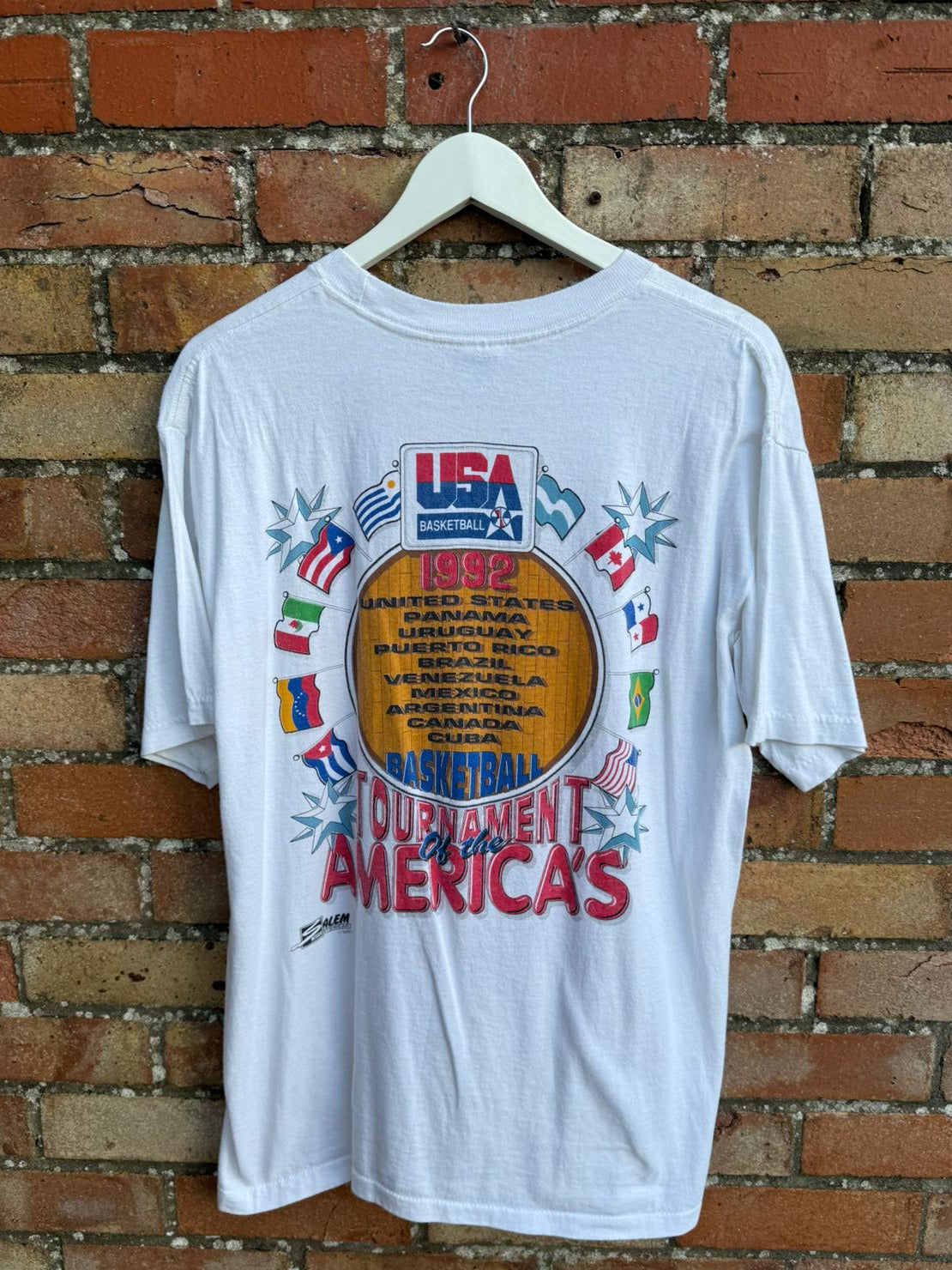 Reverse side of white t-shirt with 'USA Basketball Tournament of the Americas' design on a hanger against a brick wall.