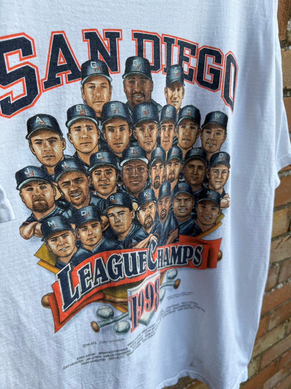 T-shirt with a graphic of baseball players and 'San Diego League Champs 1998' text, hanging against a brick wall.