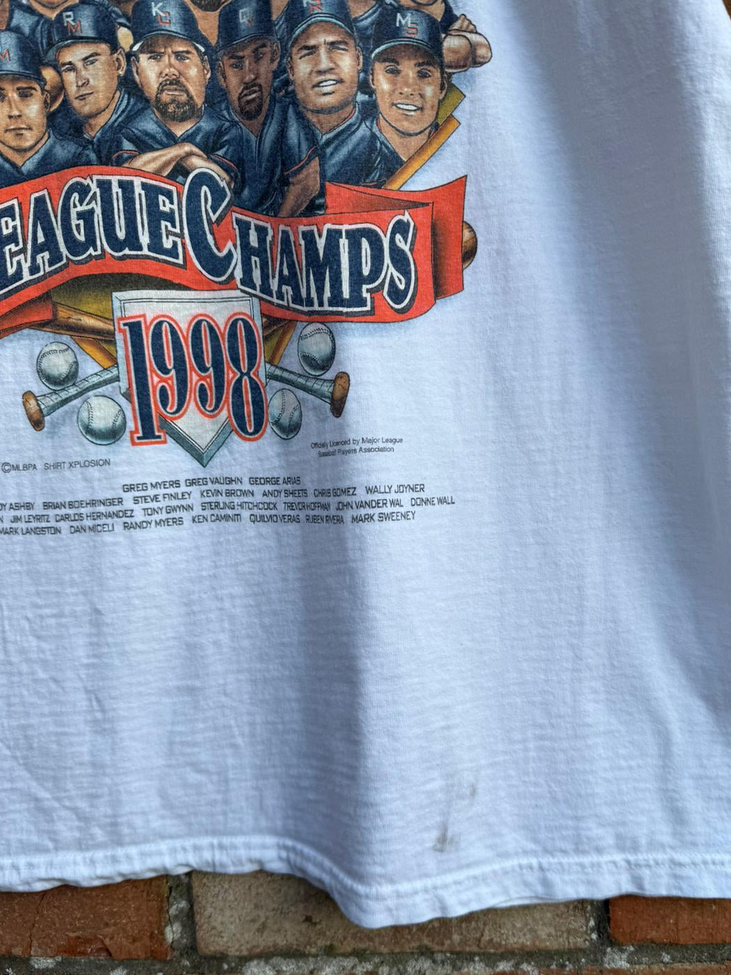 White t-shirt with 'League Champs 1998' graphic, showing a small mark on the bottom hem, hanging on a brick wall background