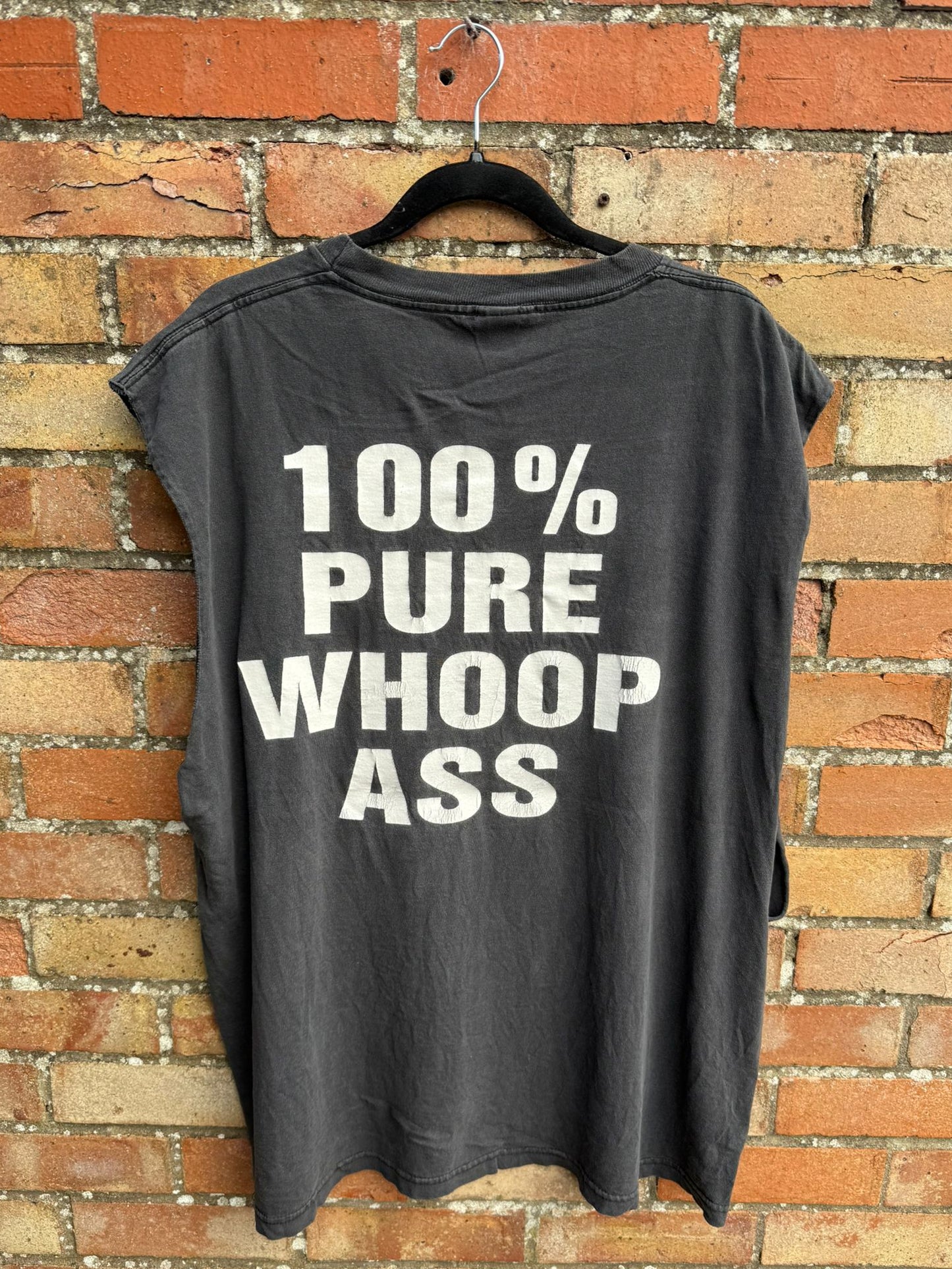 Black sleeveless shirt with white text '100% PURE WHOOP ASS' on a brick wall background