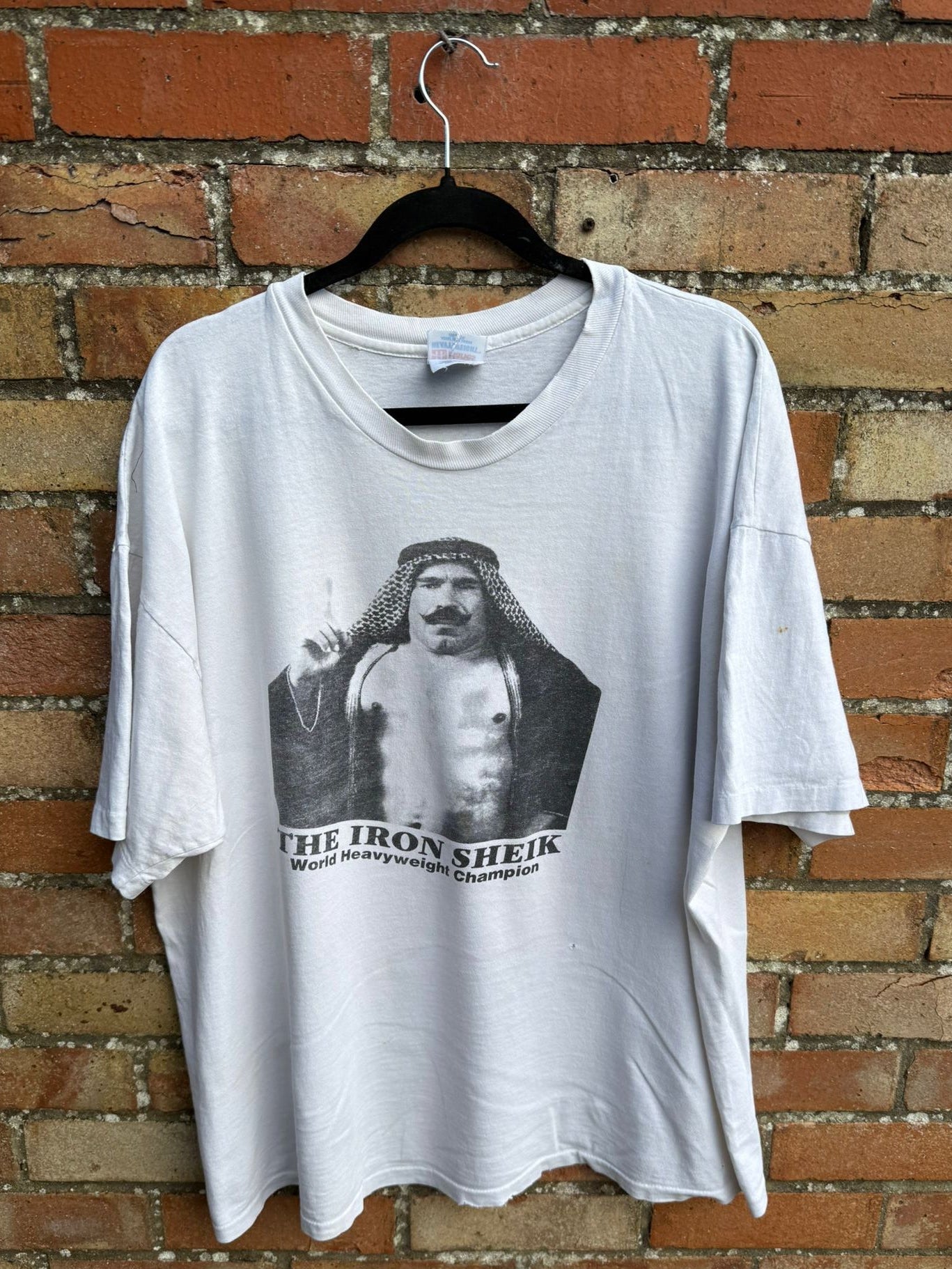 White t-shirt with 'The Iron Sheik' graphic on a hanger against a brick wall.
