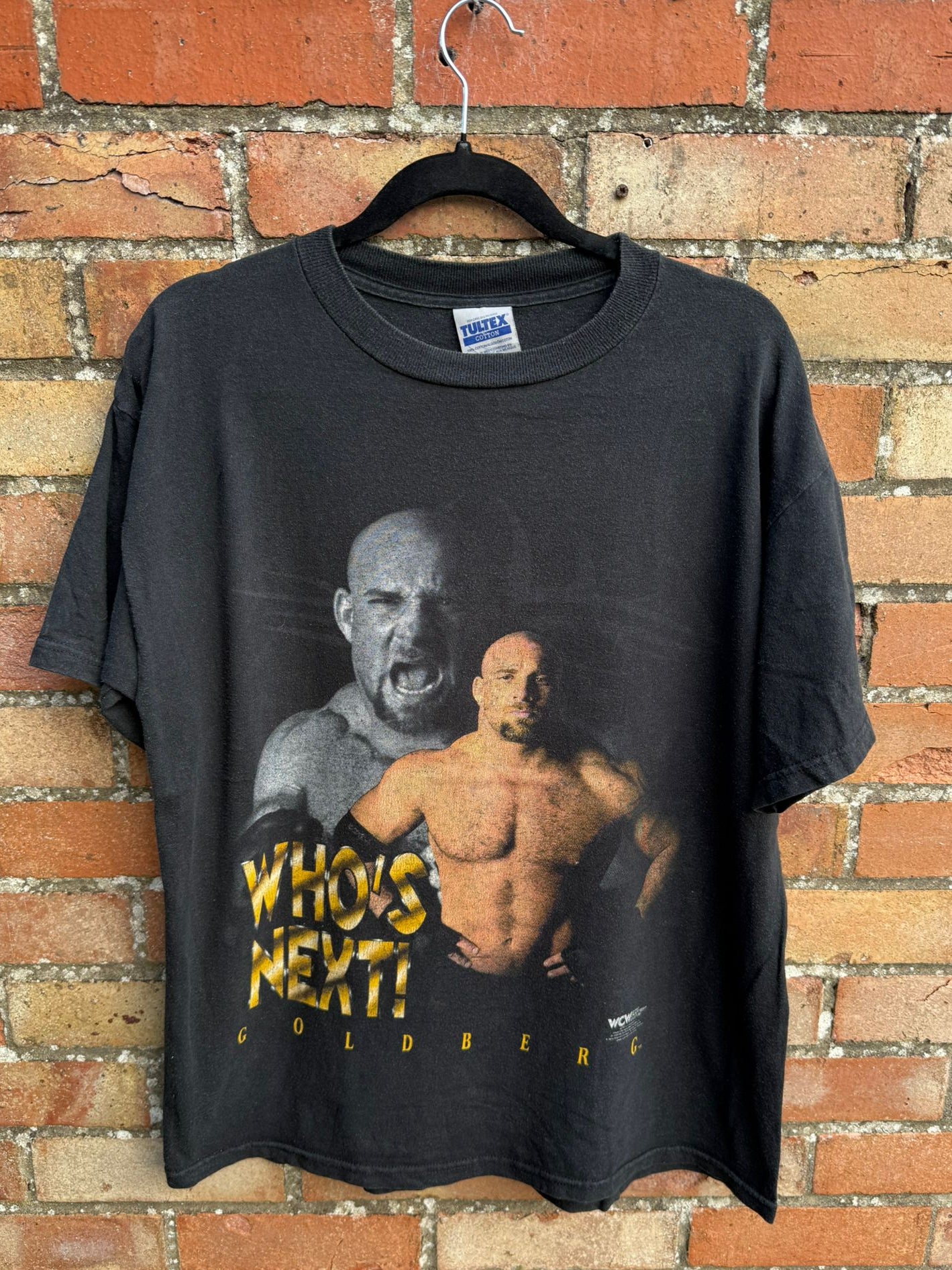 Black t-shirt with graphic design of Goldberg and text 'WHO'S NEXT!' hanging against a brick wall