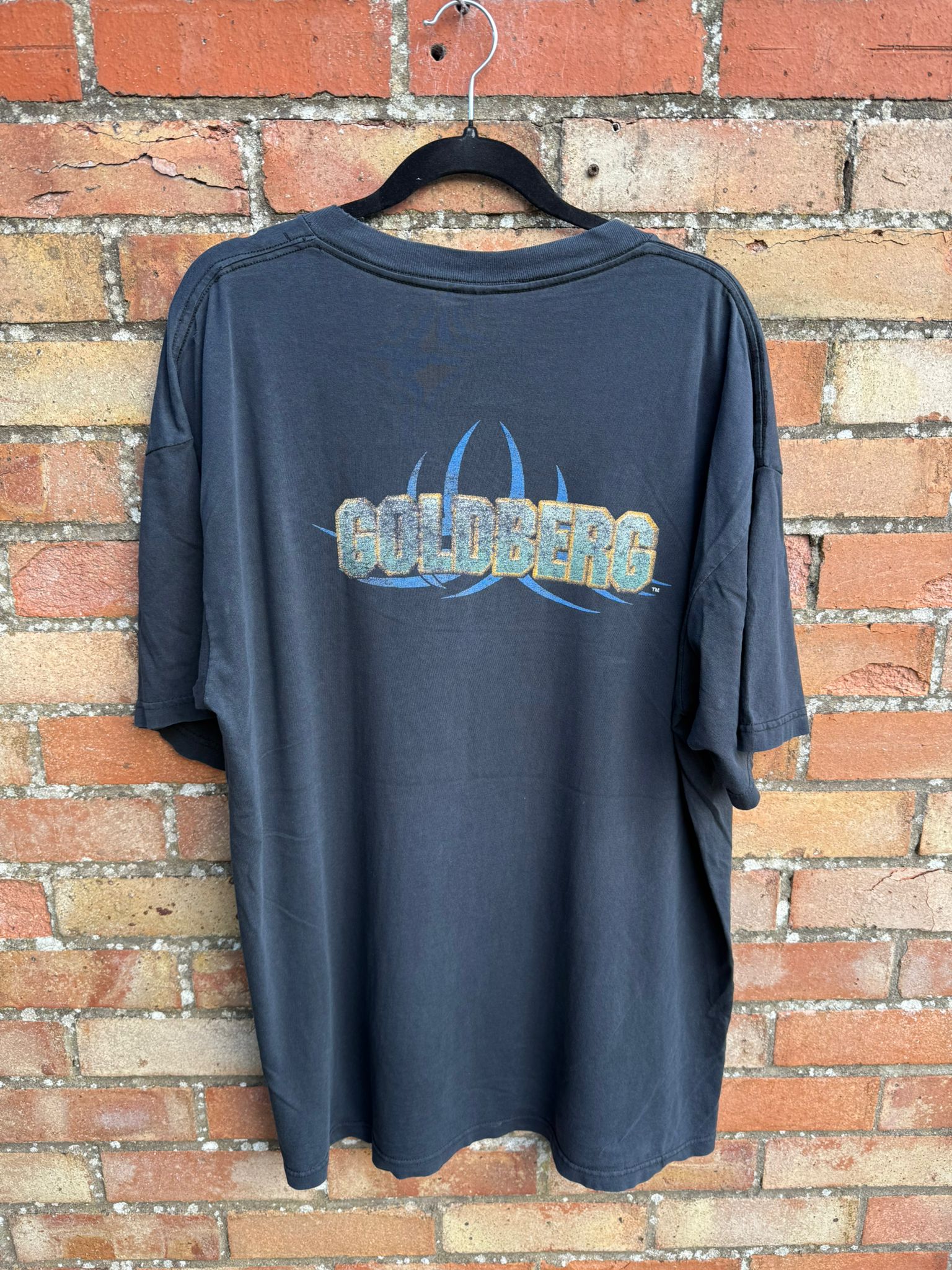 Back of a black t-shirt with Goldberg logo, hanging against a brick wall
