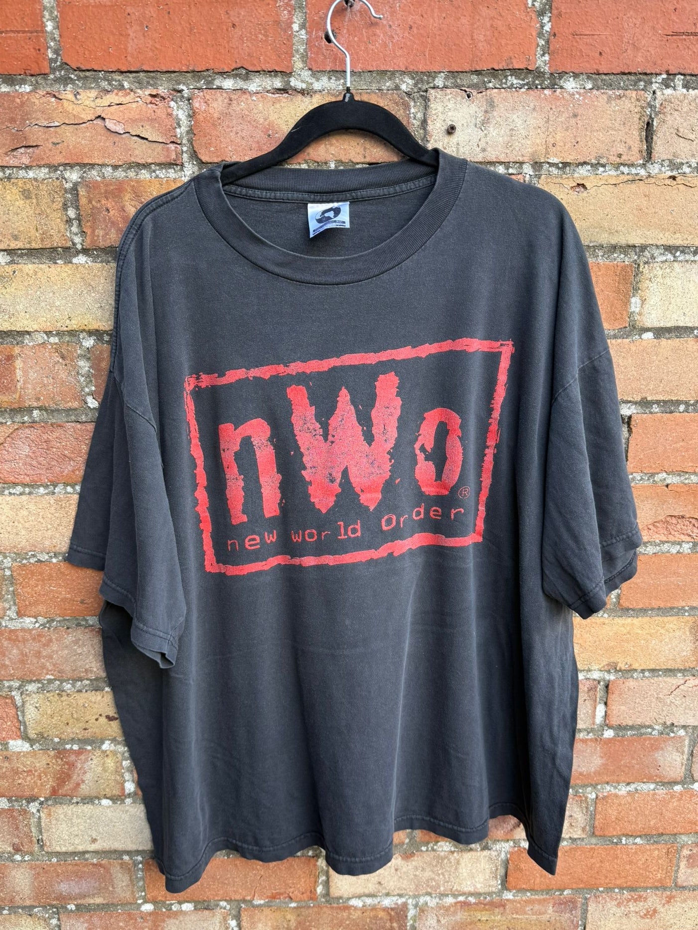 Black t-shirt with red 'nWO' new World order logo on a hanger against a brick wall.