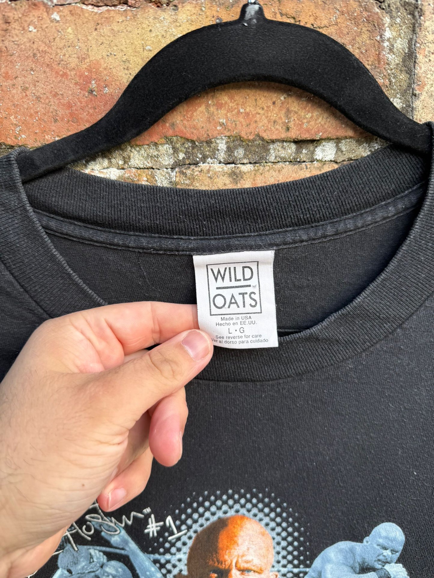 Black t-shirt with close up of 'Wild Oats' label held by a hand against a brick wall background