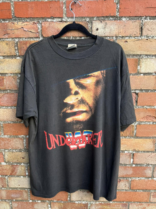 Black t-shirt with graphic design and 'Undertaker' text on a brick wall background