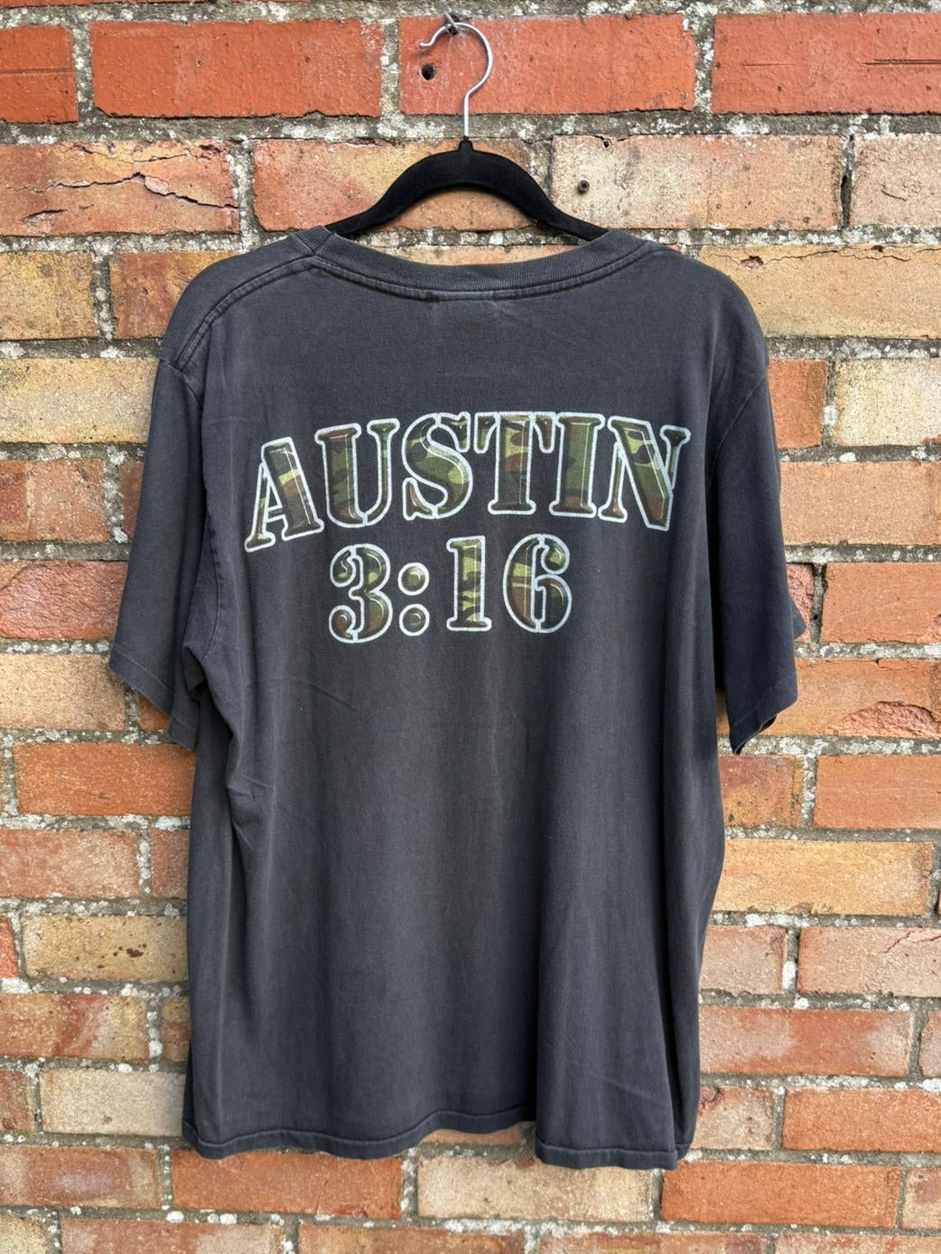 Black t-shirt with 'AUSTIN 3:16' text hanging on a brick wall background