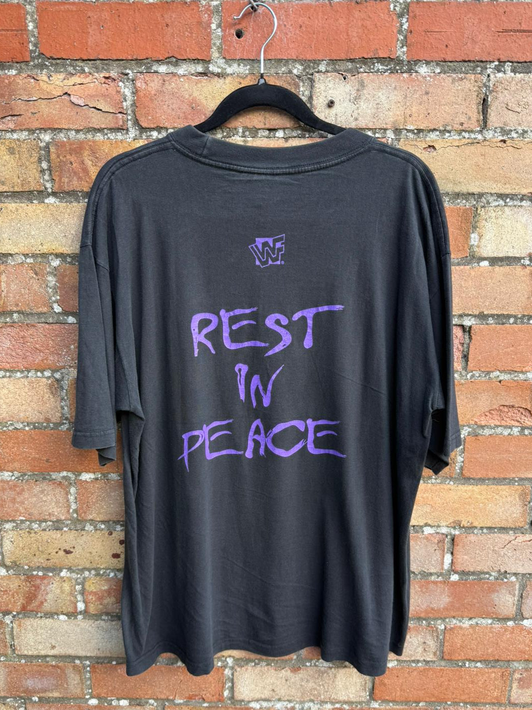 Back of black t-shirt with 'REST IN PEACE' text and WWF logo on a hanger against a brick wall.