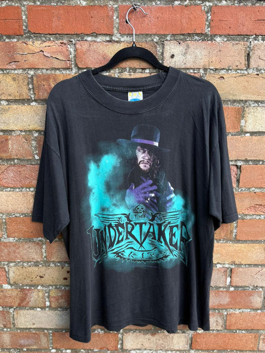 Black t-shirt with graphic design of a person in a top hat and 'Undertaker' text, hanging against a brick wall.