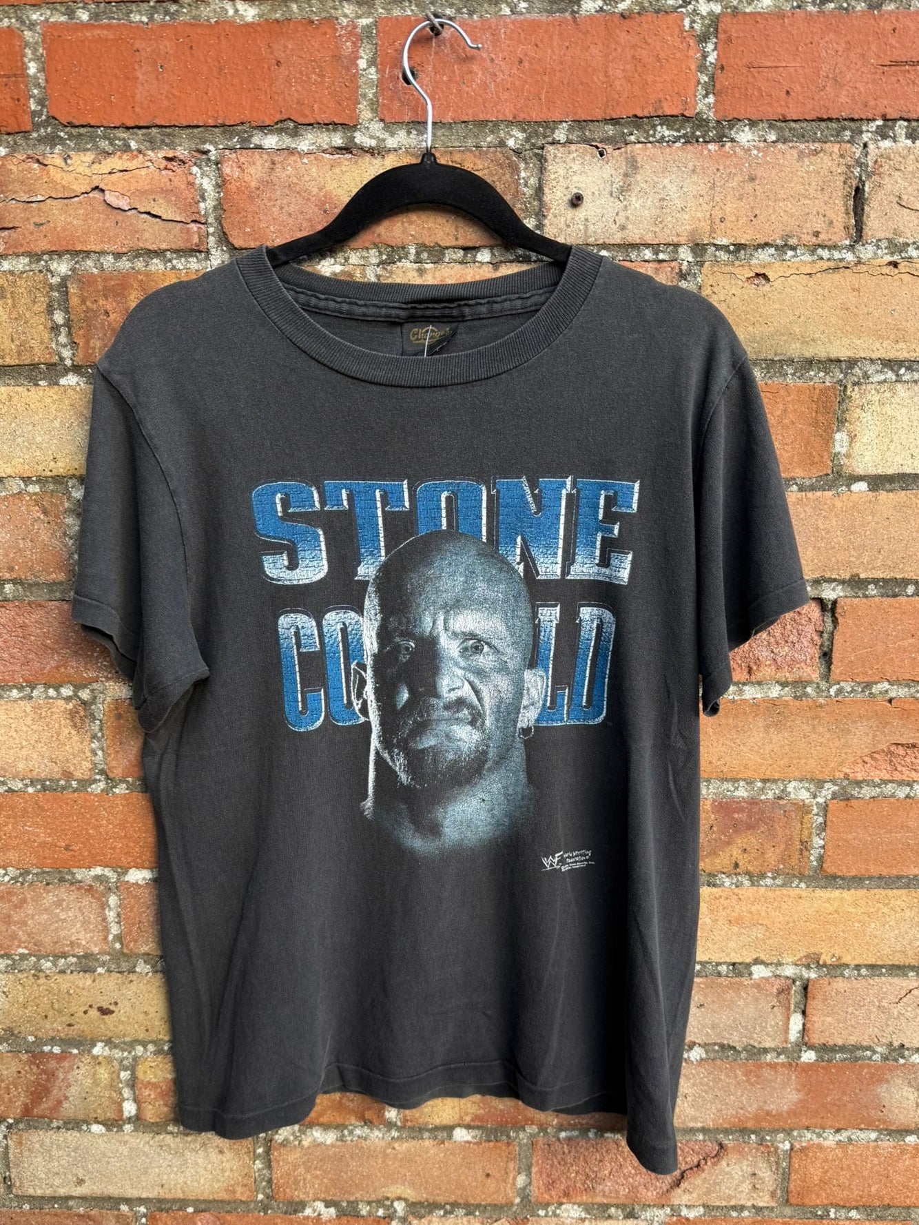 Black t-shirt with text STONE COLD and graphic design hanging on a brick wall.