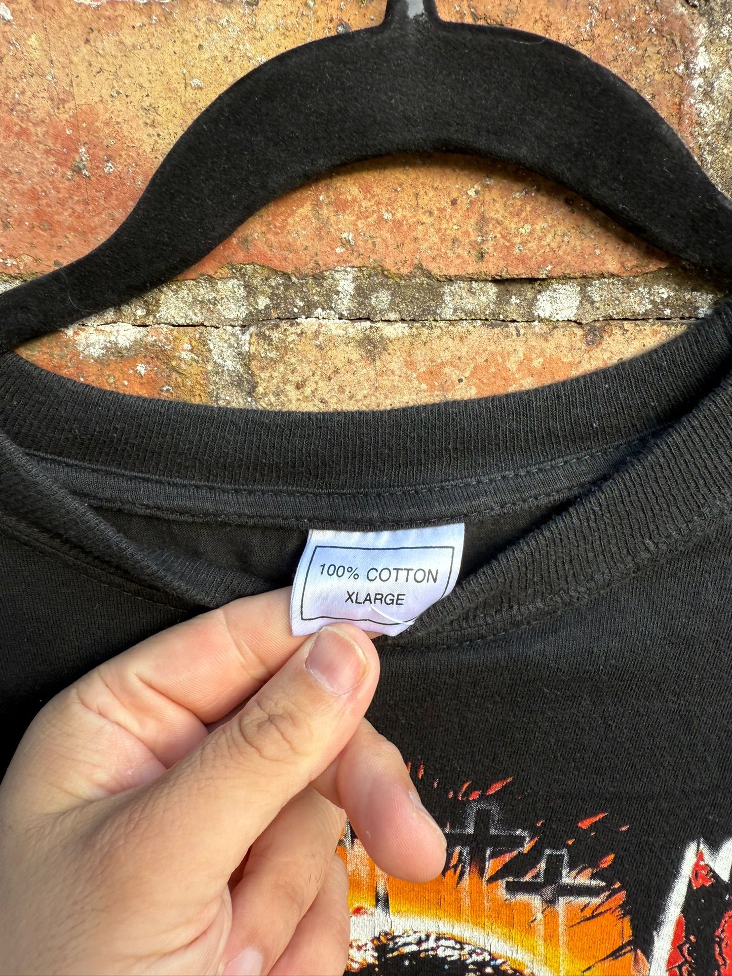 Black t-shirt with a label showing '100% Cotton XLarge' held by a hand against a brick wall.