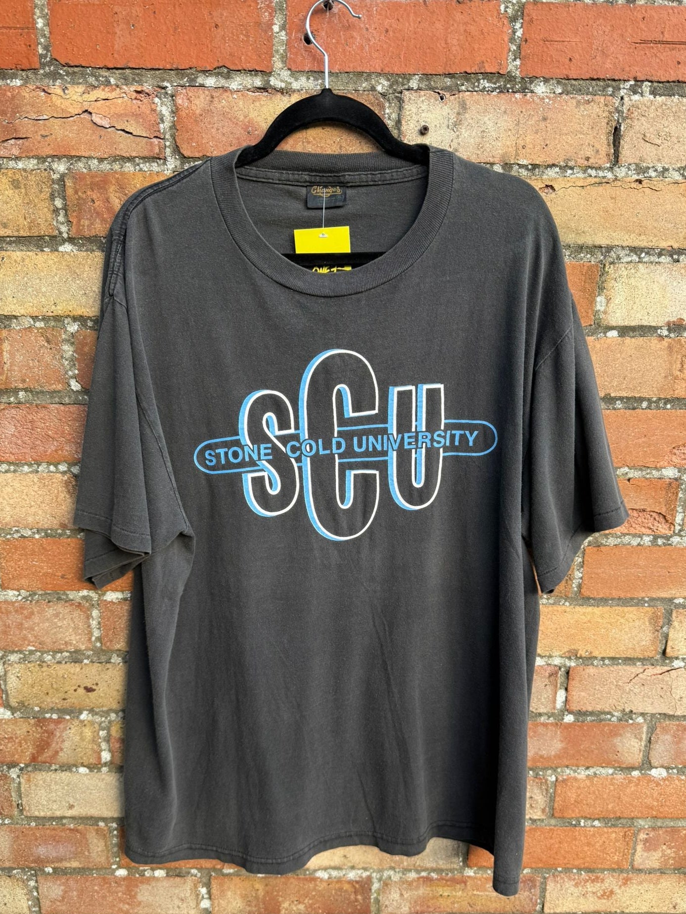 Black t-shirt with 'SCU' Stone Cold University logo on a hanger against a brick wall