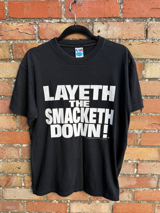Black t-shirt with white text 'LAYETH THE SMACKETH DOWN!' on a hanger against a brick wall.