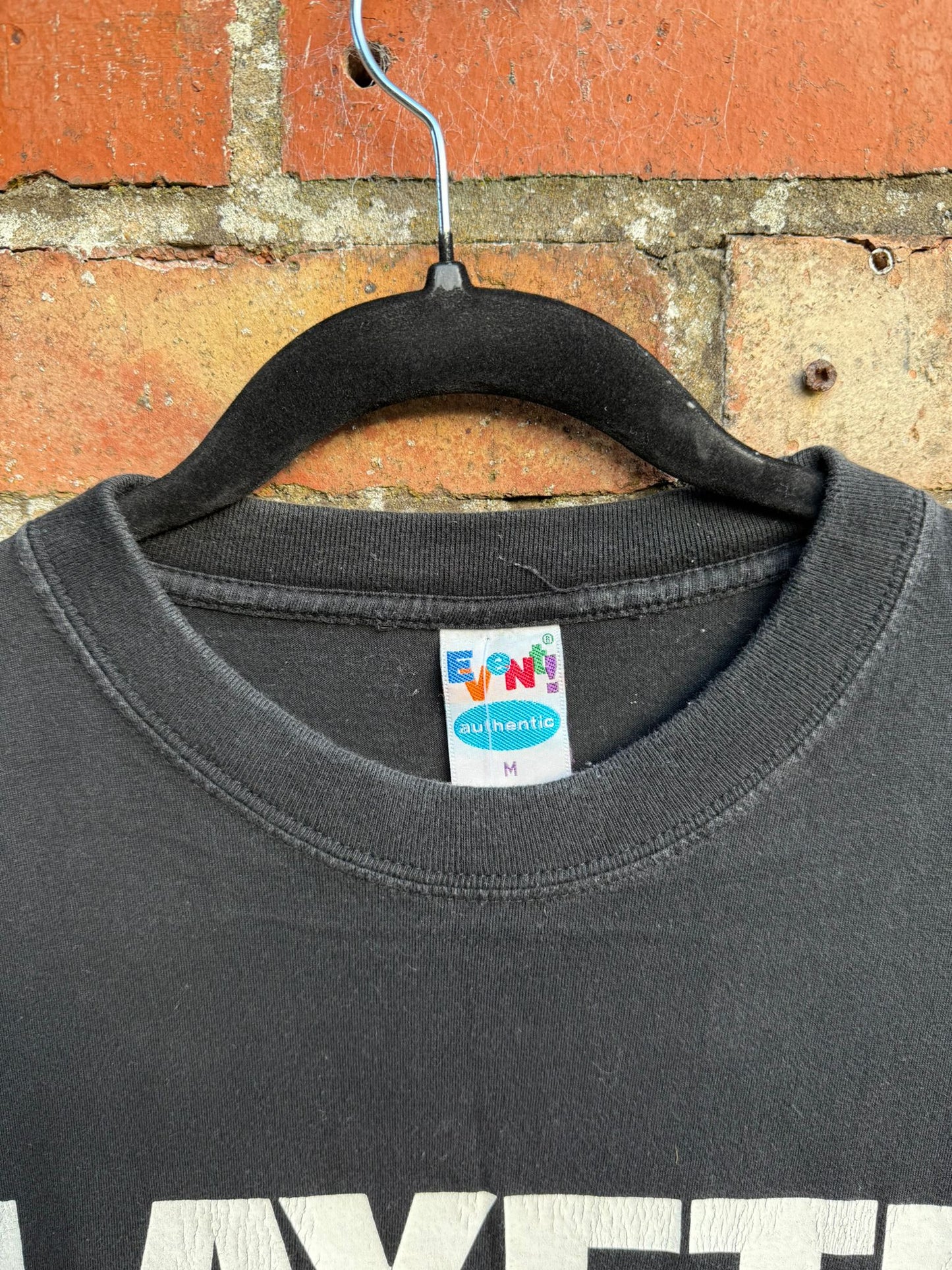 Black t-shirt with a visible Event brand label on a hanger against a brick wall.