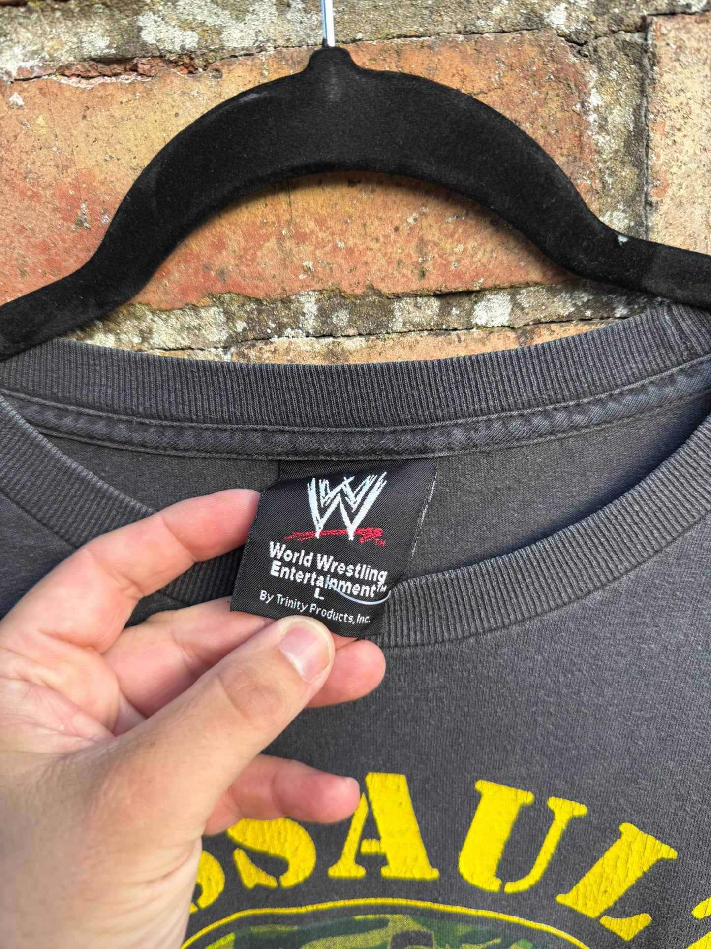 Black t-shirt with yellow text and WWE logo tag, hanging against a brick wall.