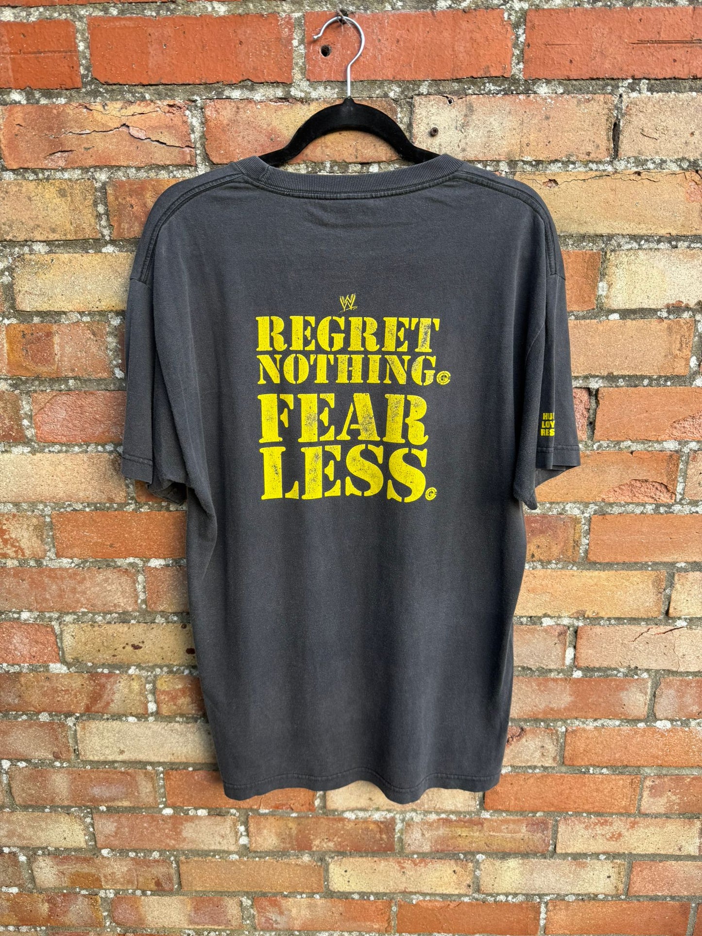 Black t-shirt with yellow text 'REGRET NOTHING. FEAR LESS.' on a brick wall background