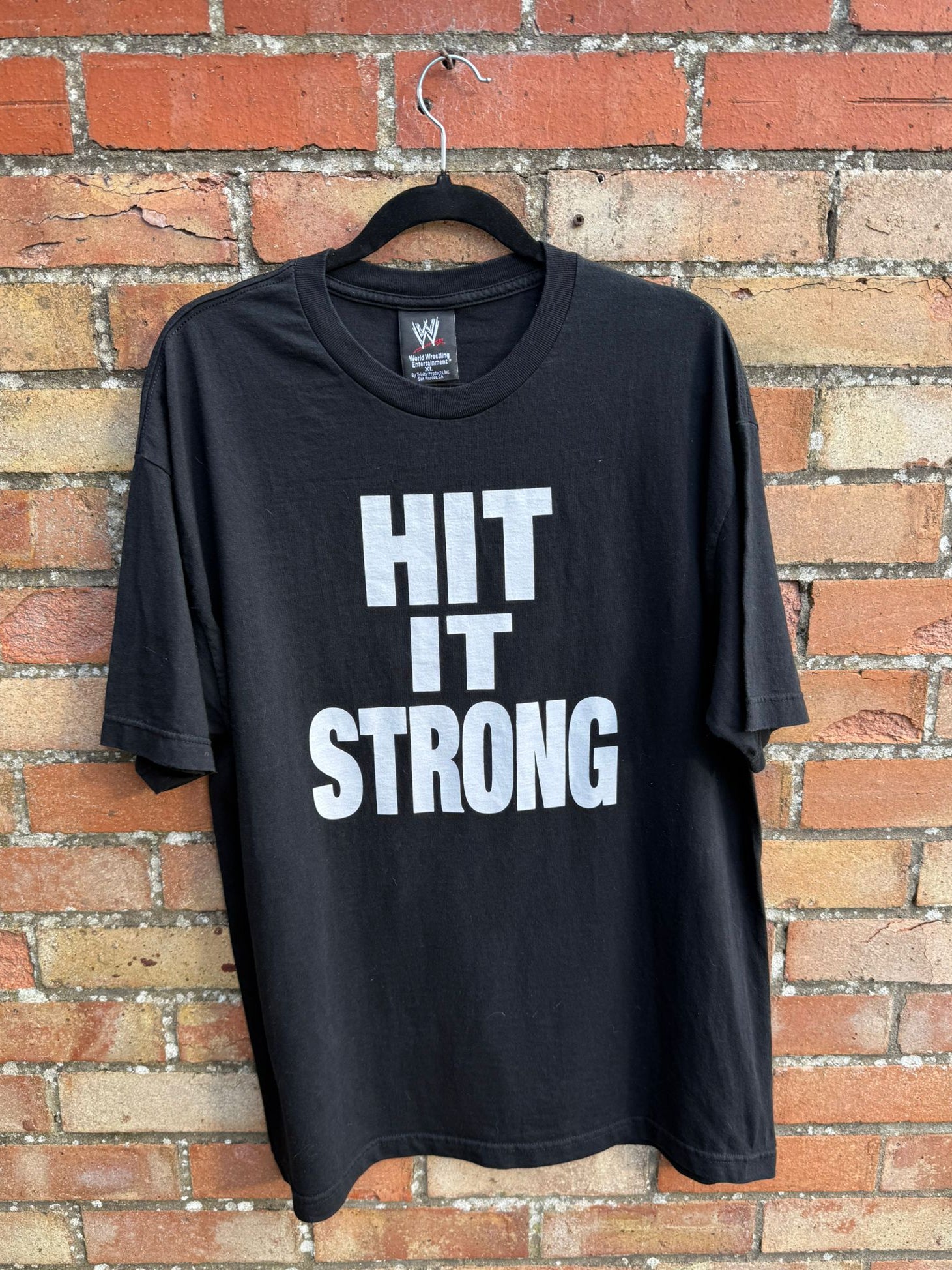 Black t-shirt with white large print Hit it Strong hanging against a brick wall