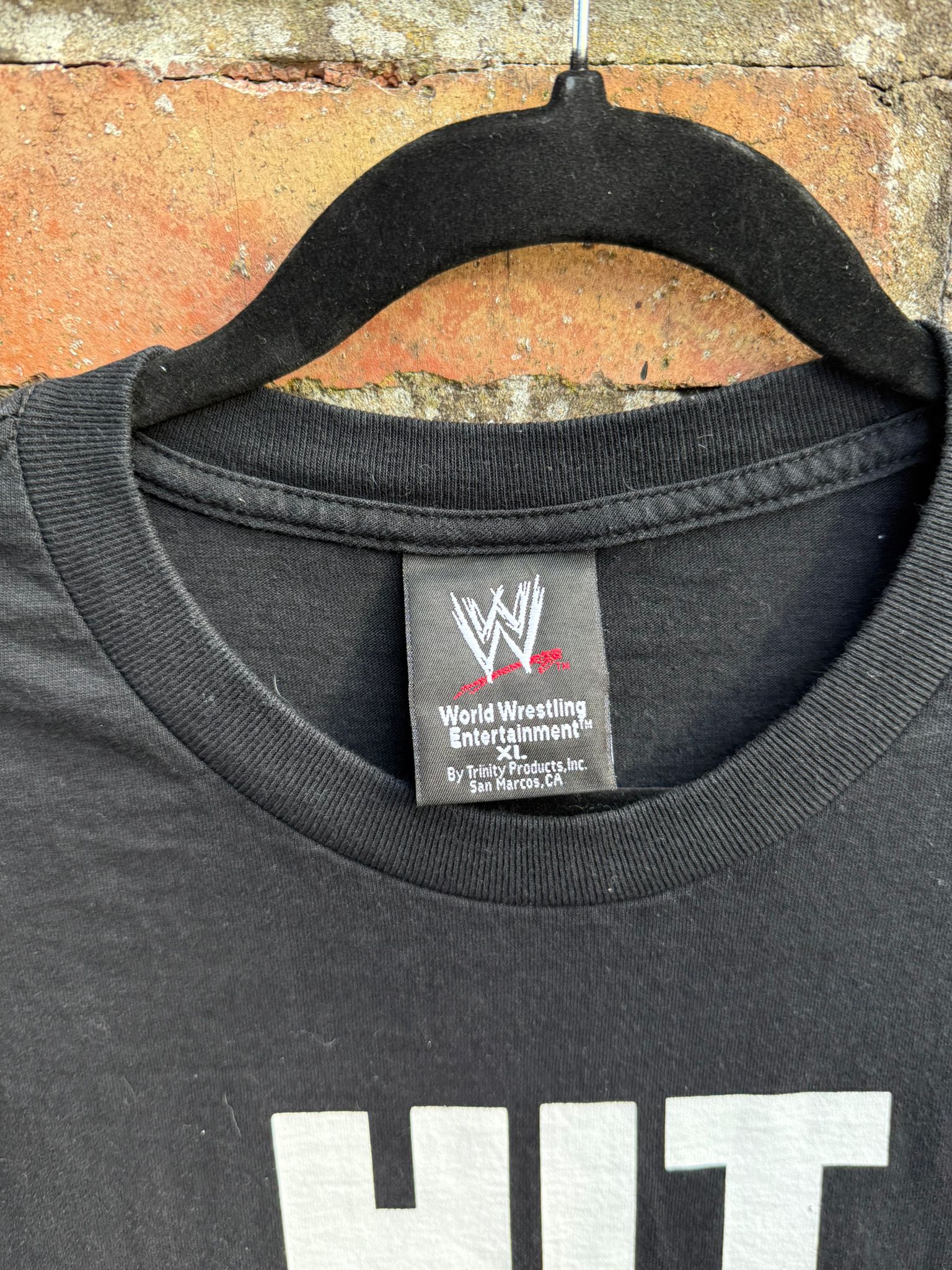Black t-shirt with WWE logo tag on a hanger against a brick wall.