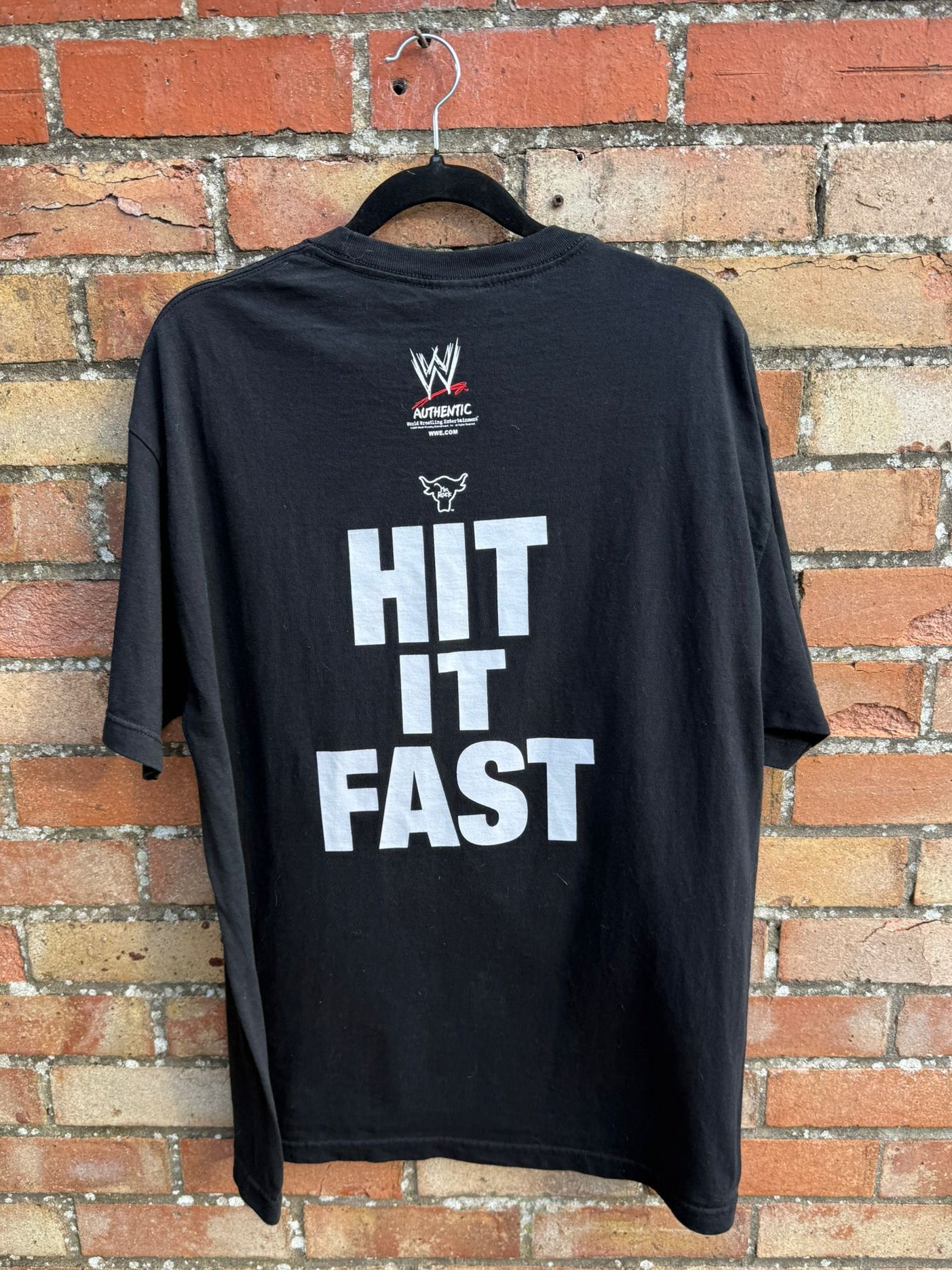Black t-shirt with WWE logo and 'HIT IT FAST' text on a hanger against a brick wall.