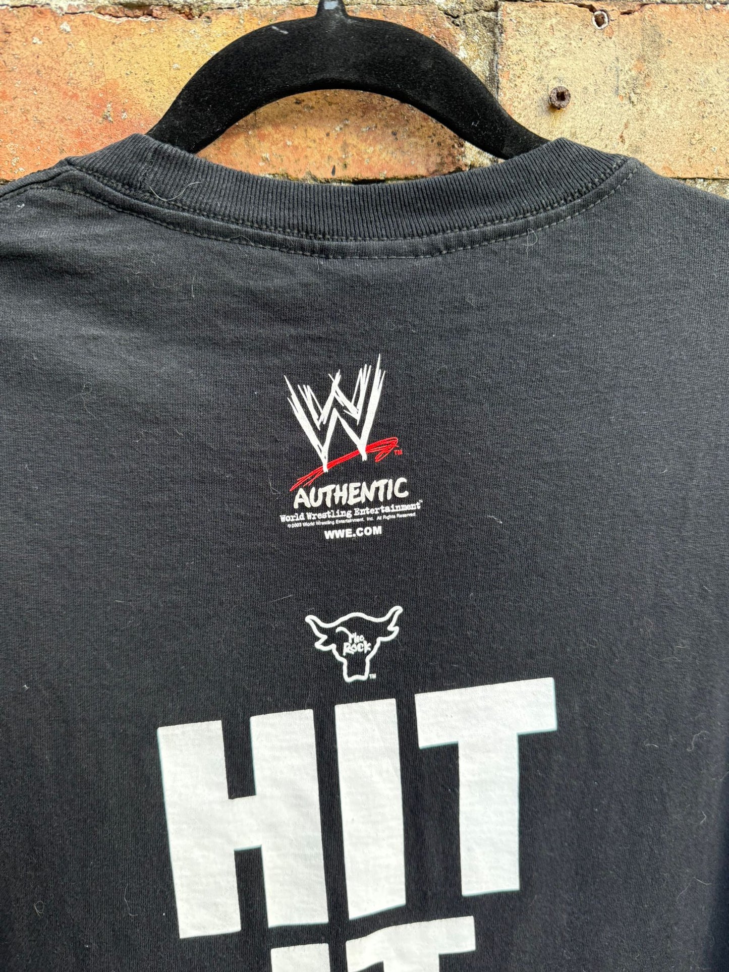 Black t-shirt with WWE logo and text on a hanger against a brick wall.