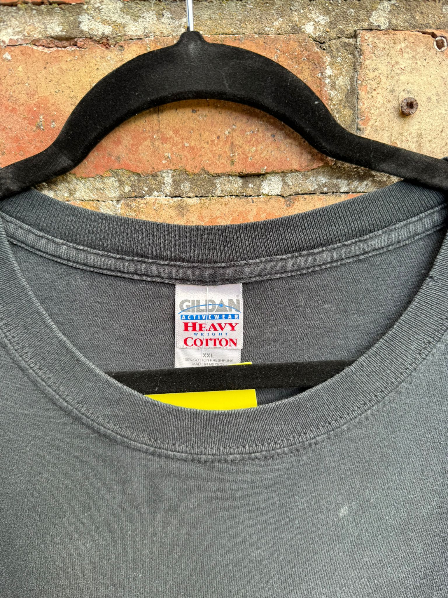 Close-up of black t-shirt with a Gildan Heavy Cotton label on a hanger against a brick wall.