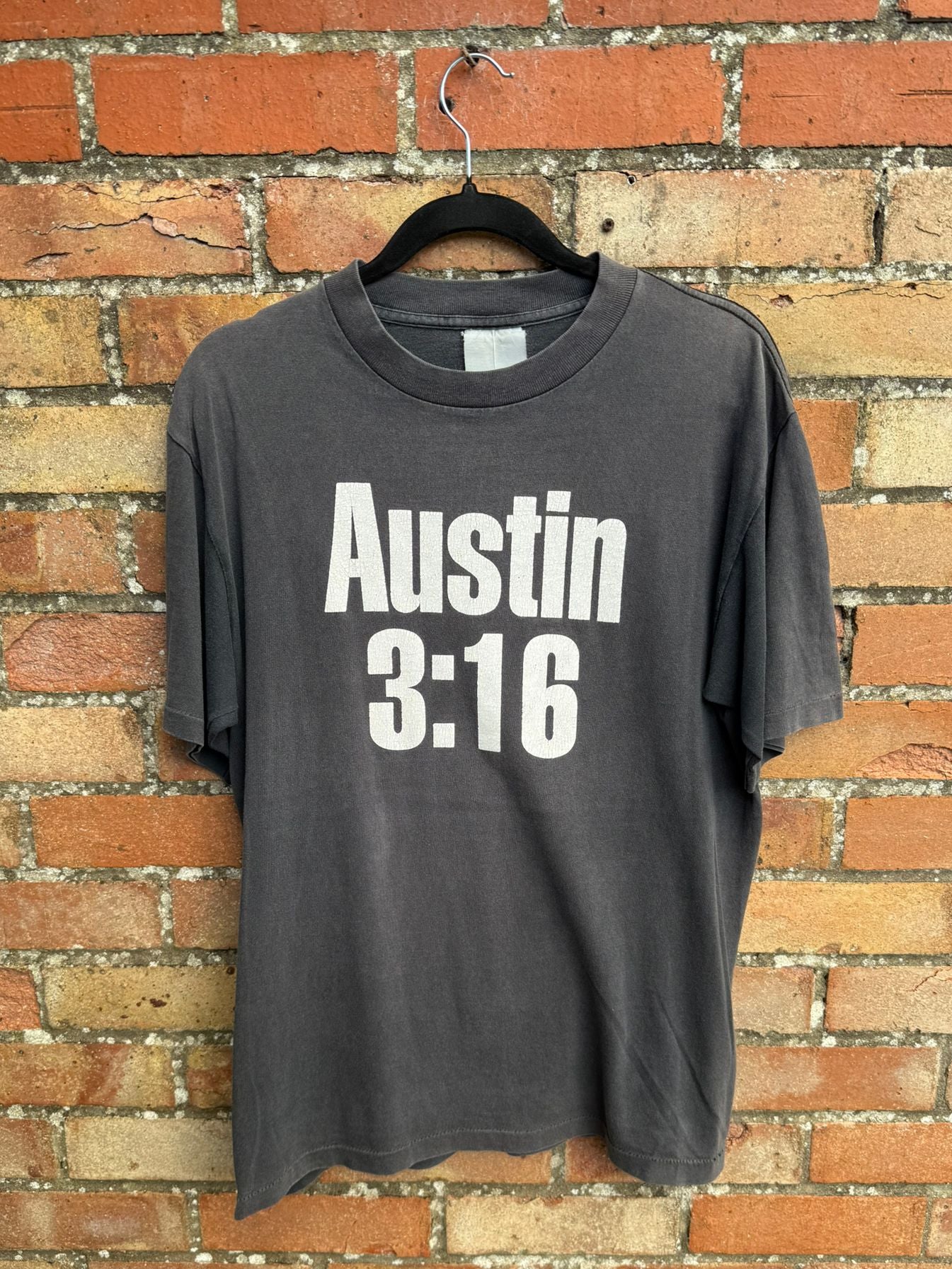 Black t-shirt with 'Austin 3:16' text, hanging against a brick wall background