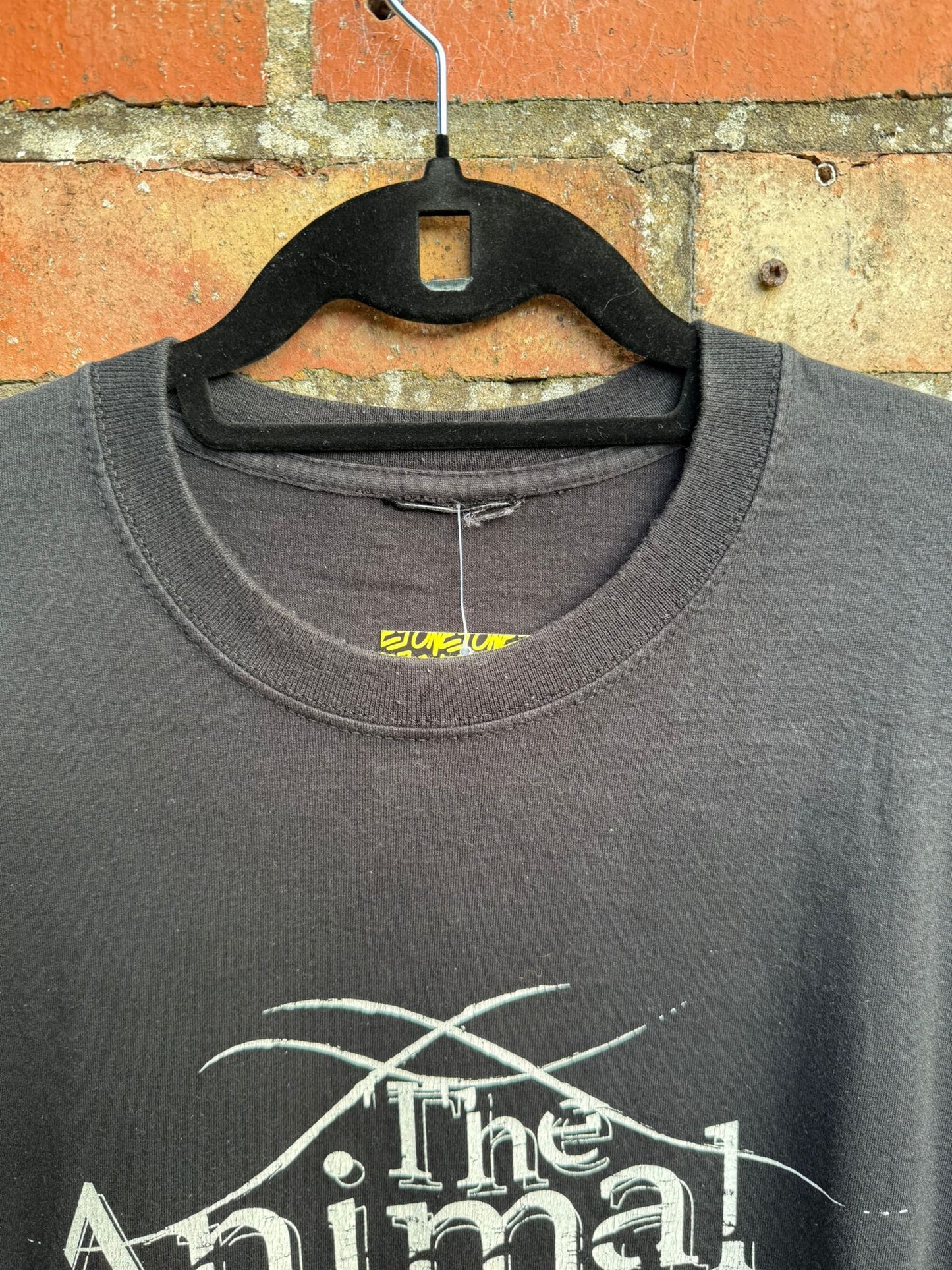 Black t-shirt with 'The Animal' text and showing a cut tag, on a hanger against a brick wall.